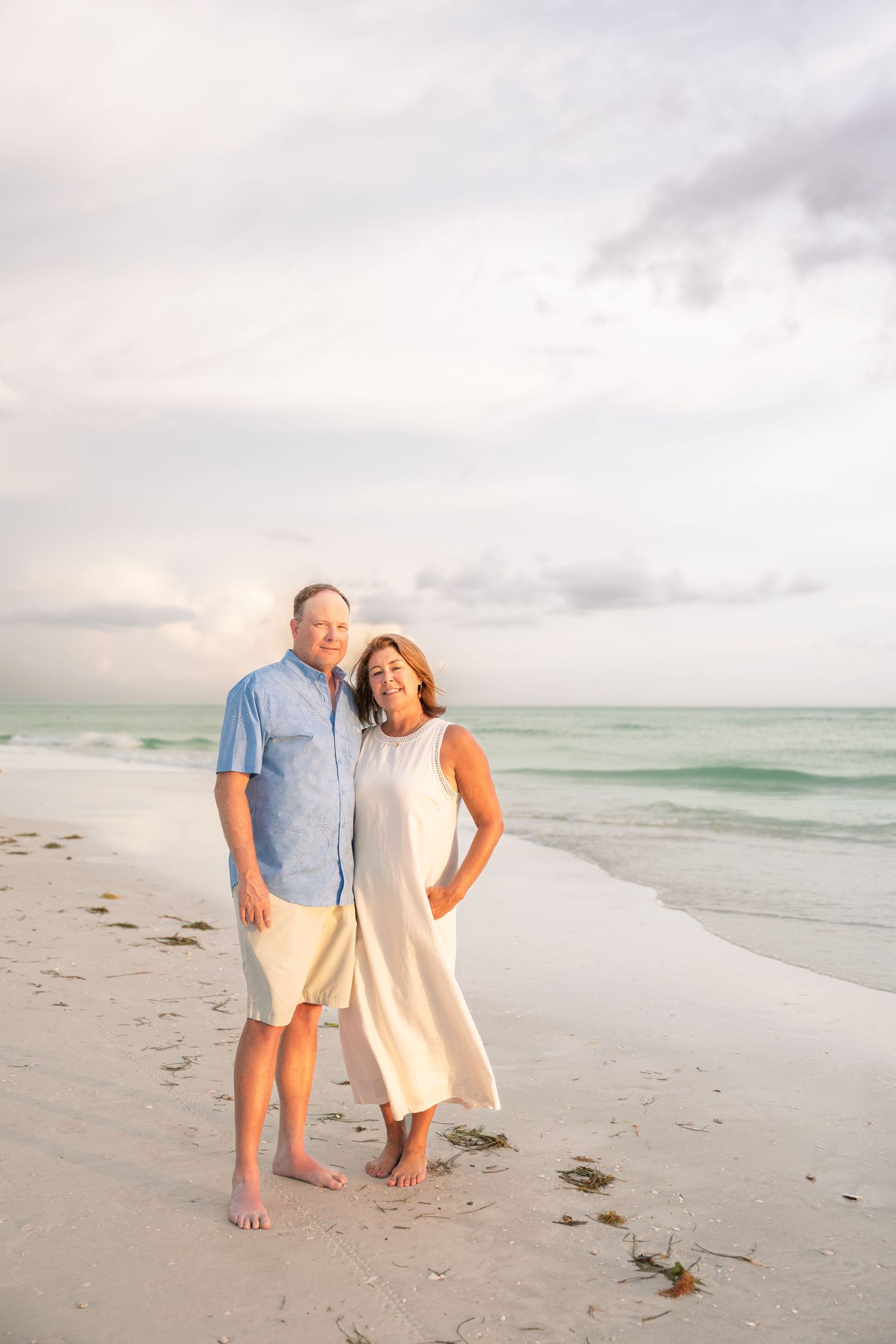 A couple smiles together on a white sand beach at sunset, man in blue shirt and woman in white dress with turquoise ocean behind.