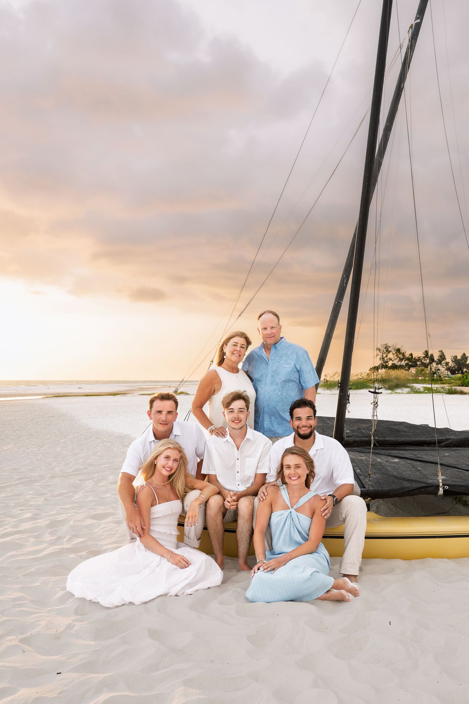 Family of seven posing together on a sandy beach at sunset near a sailboat, dressed in white and light blue outfits.