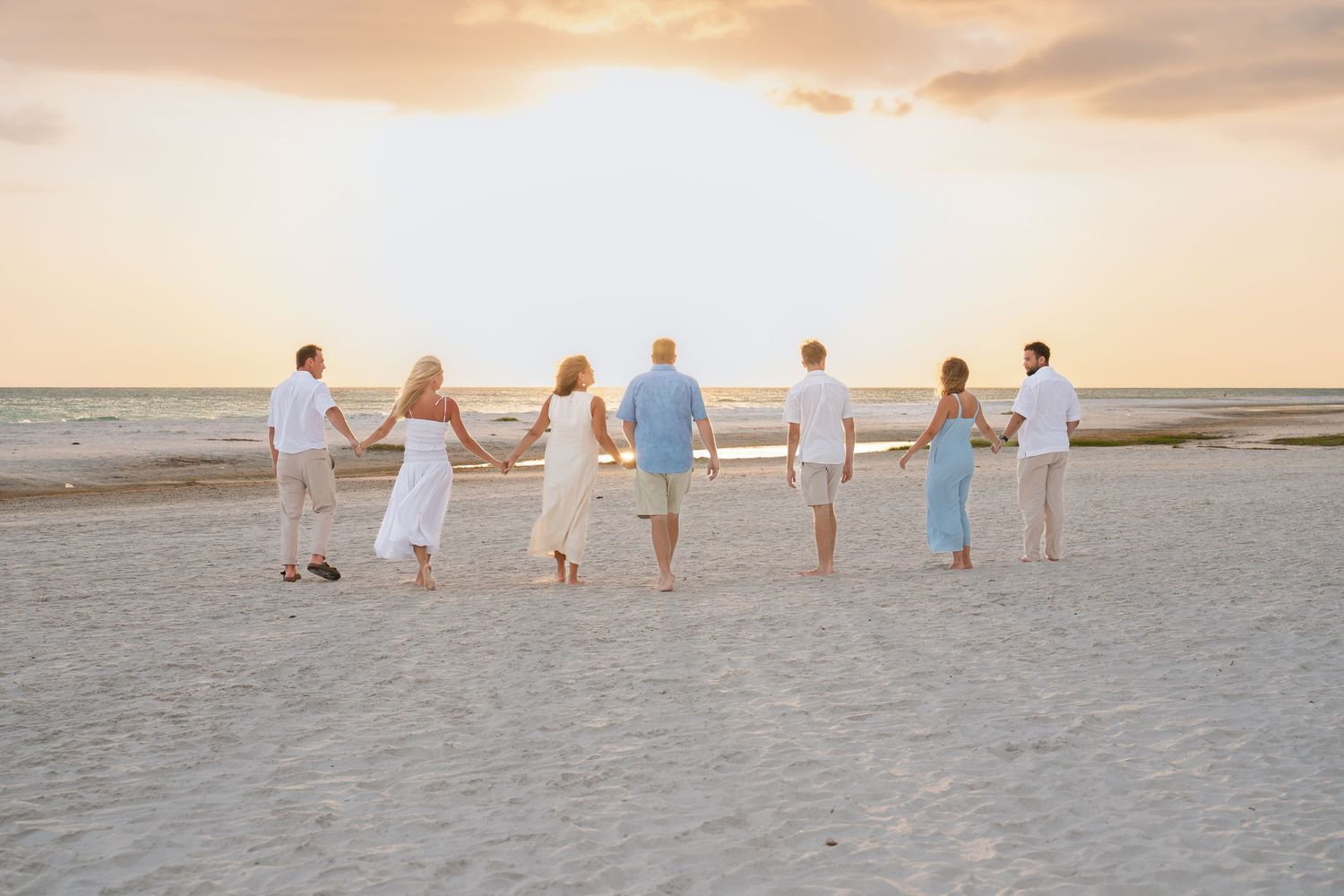 A family holds hands walking toward a stunning beach sunset, dressed in coordinating white and blue outfits.