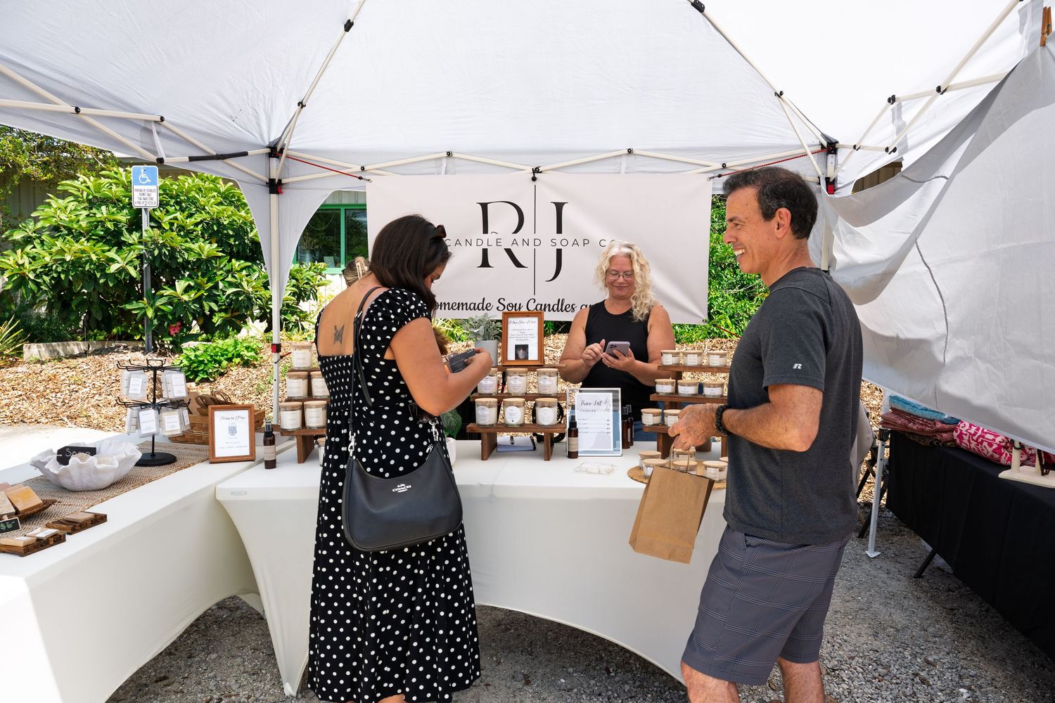 Customers browse handmade goods at an outdoor farmers market vendor booth under a white canopy tent.