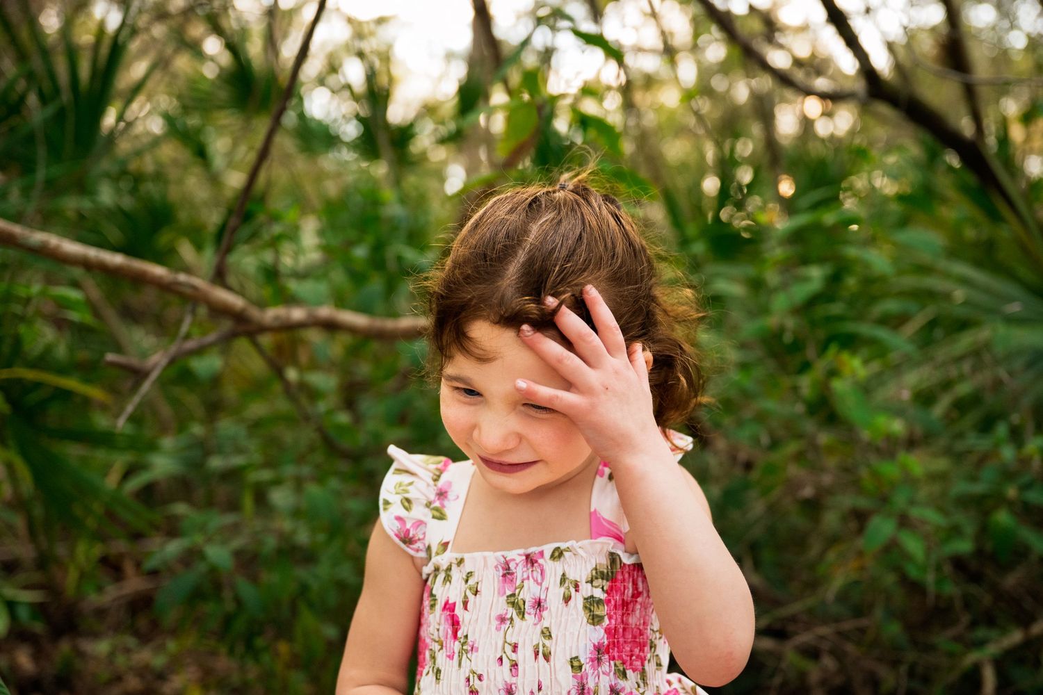 A person in a floral dress stands in a lush garden setting with sunlight filtering through the trees.