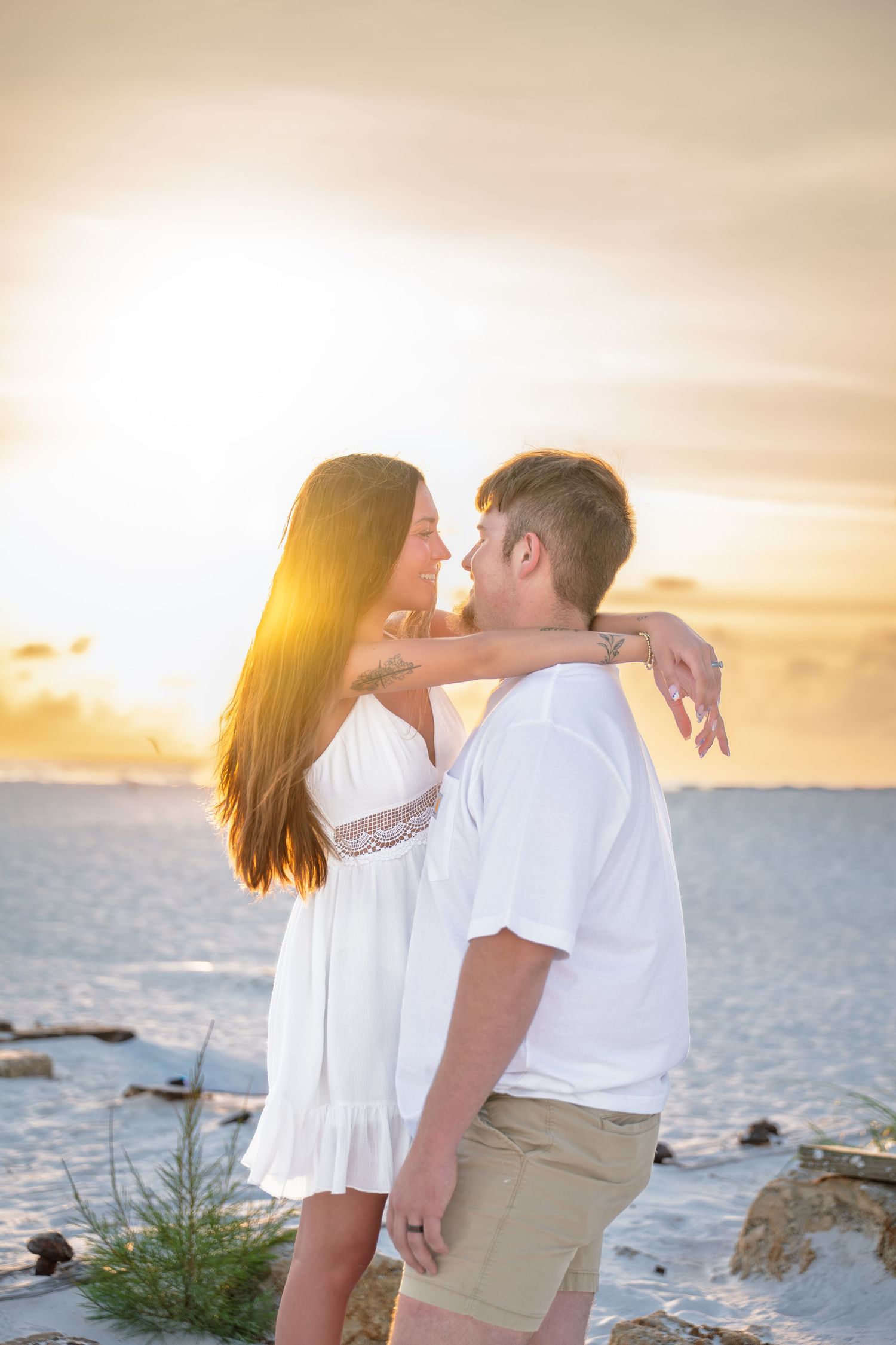 Romantic couple embracing on a white sand beach at golden sunset, woman in white dress, man in white shirt.