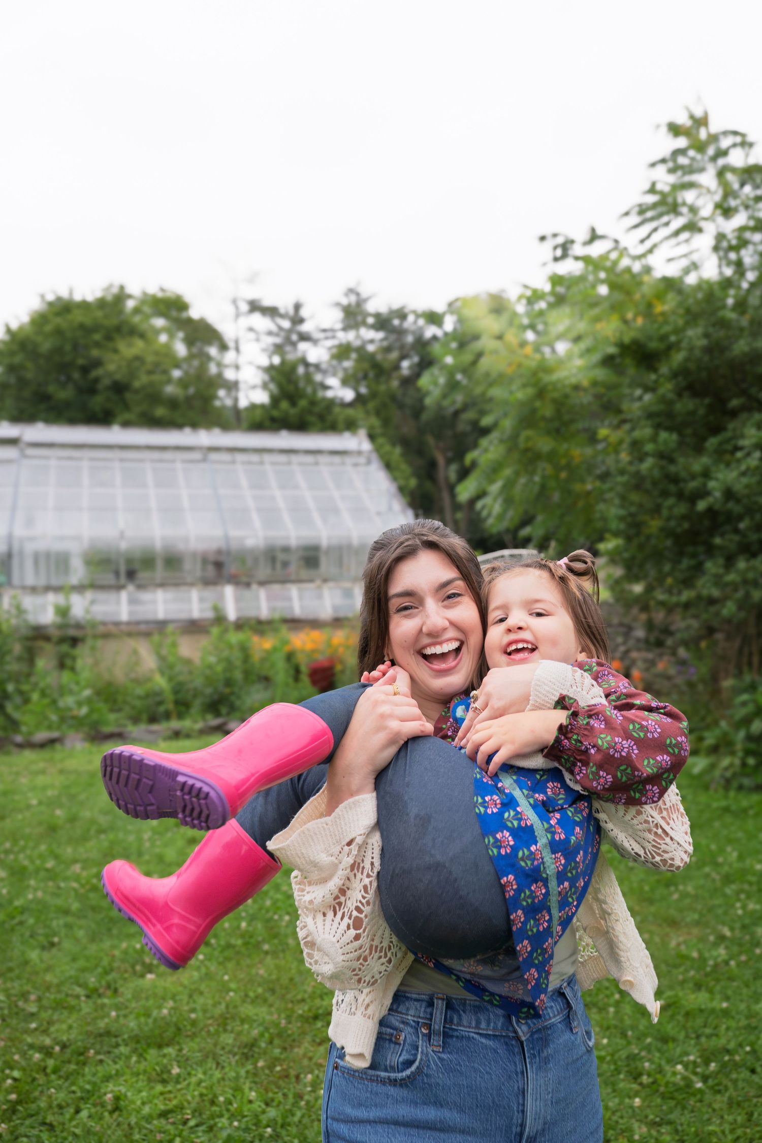 A joyful woman carries a laughing little girl in pink rain boots outdoors near a greenhouse garden.