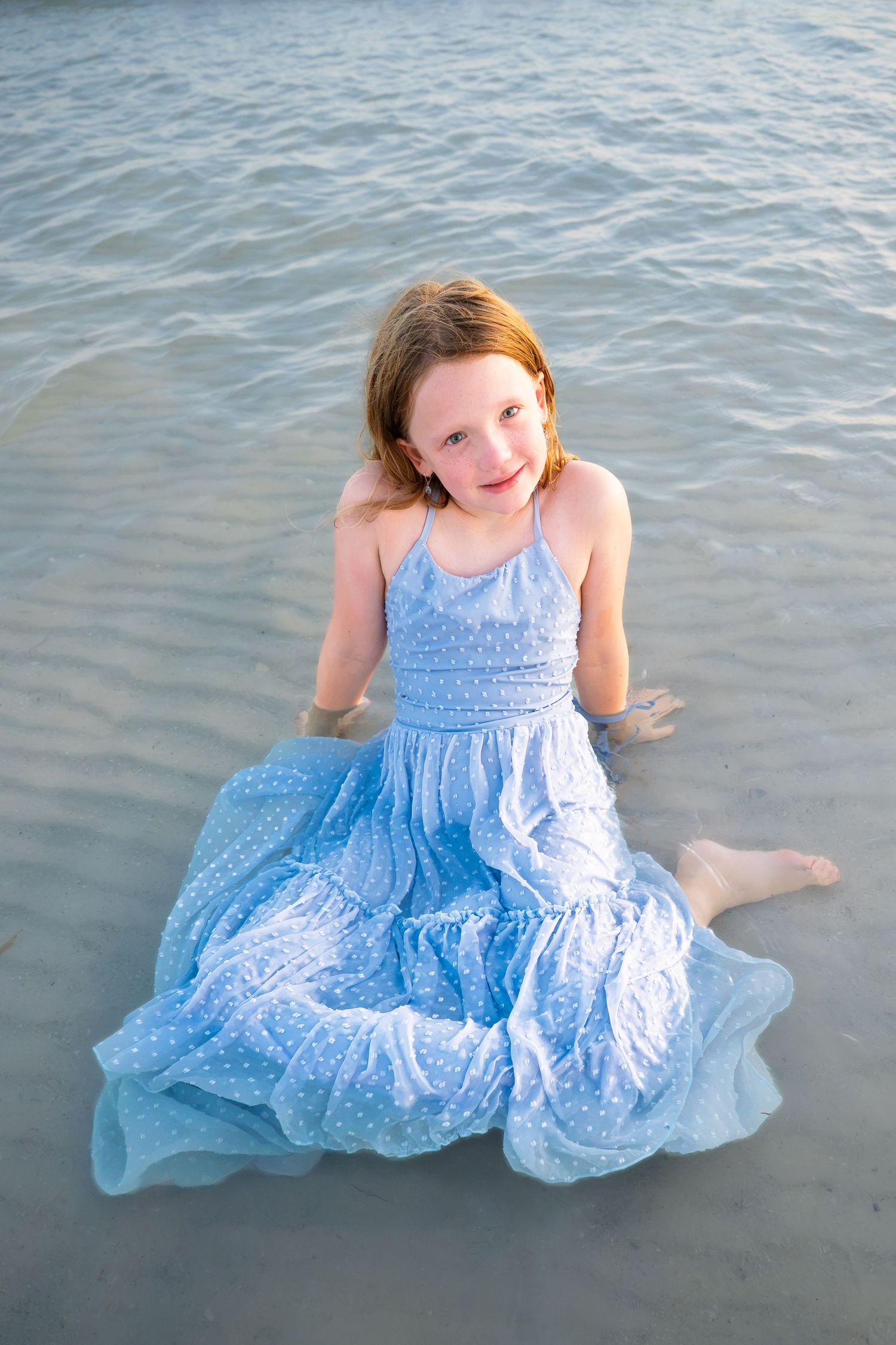 Young girl in blue polka dot dress sitting on sandy beach shore, smiling at camera with ocean water behind her.