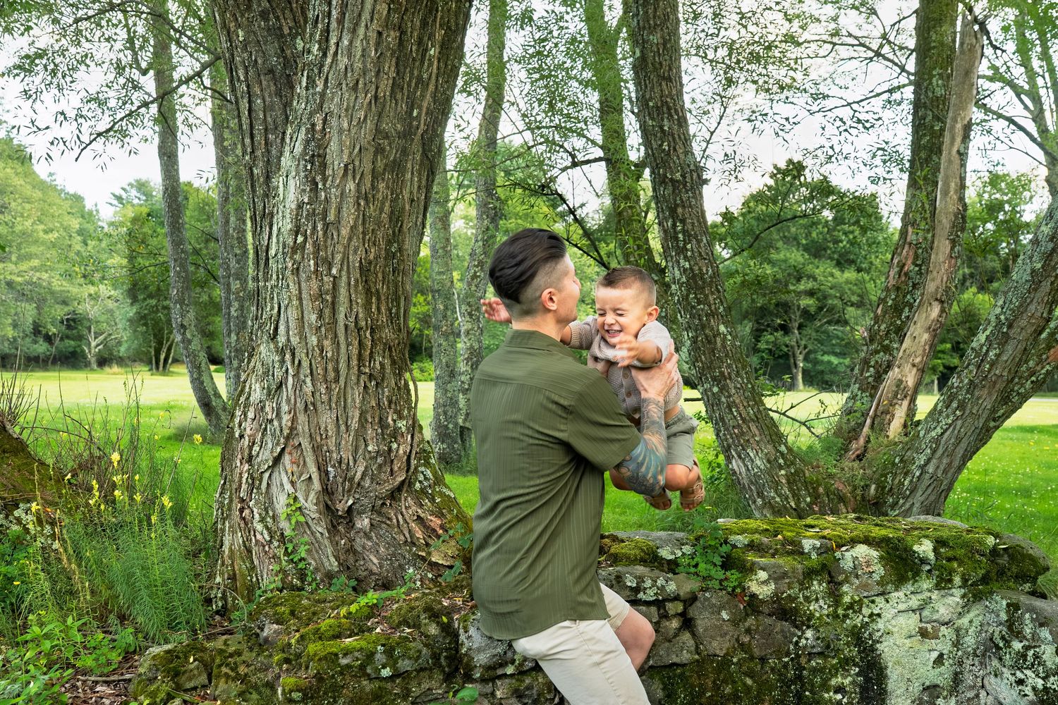 Father holding smiling baby outdoors near mossy stone wall and trees in lush green park setting.