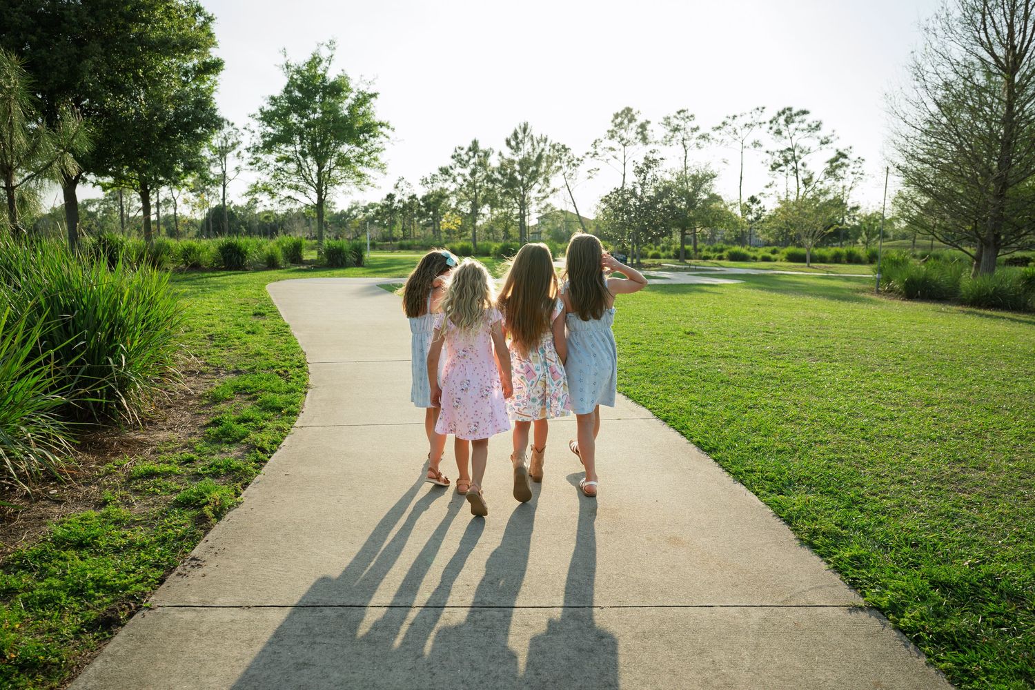 Four girls walking hand in hand down a sunny park pathway surrounded by lush green grass and trees.
