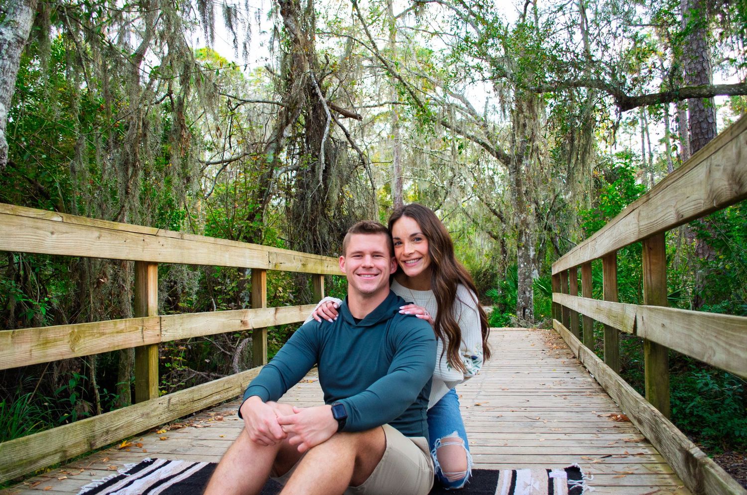 A wooden boardwalk path winds through a lush green forest with Spanish moss hanging from trees.