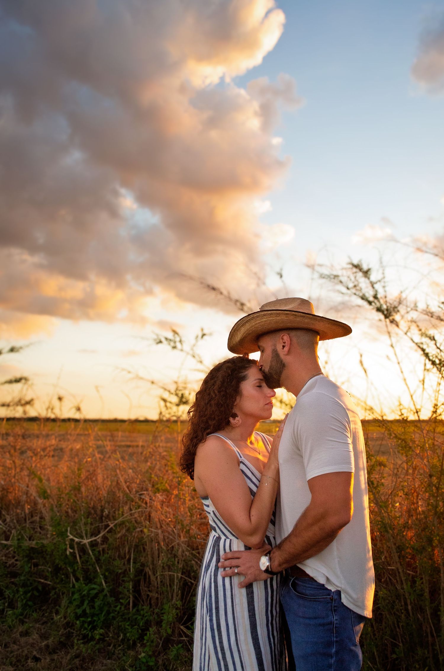 A couple embraces in a golden field during sunset as warm clouds glow overhead in a romantic outdoor photoshoot.