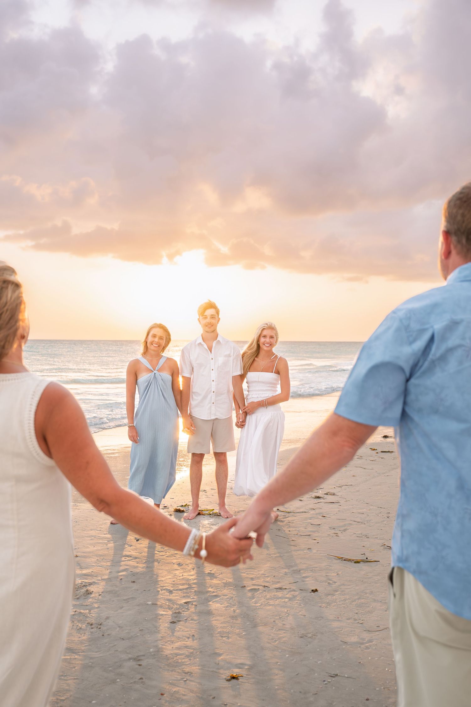 Family holding hands on a beach at sunset, with two young adults smiling in the background by the ocean.