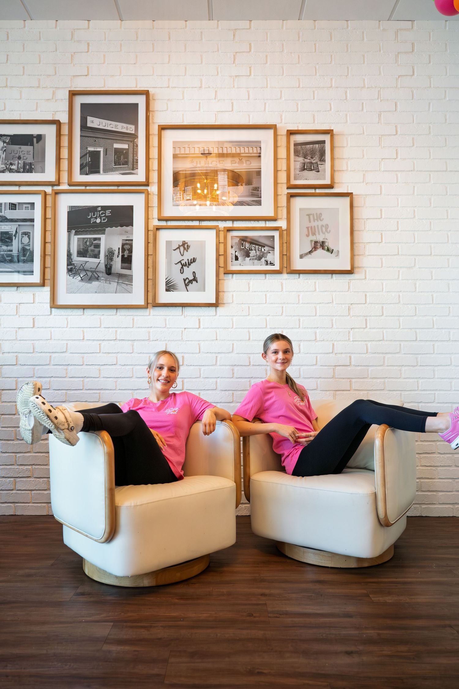 Two women in pink shirts relax in white swivel chairs against a white brick wall with framed black and white photos.