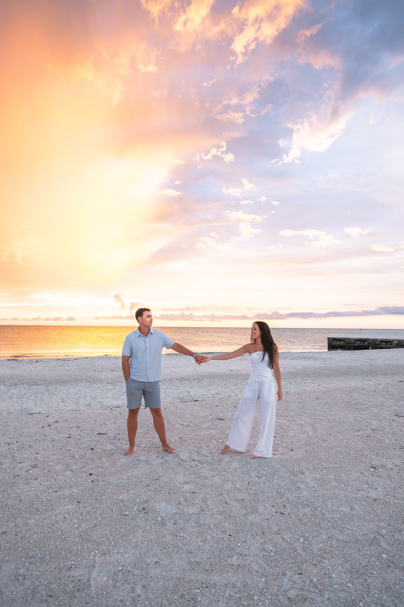 Couple holding hands on a sandy beach at sunset with colorful pink and orange clouds over the ocean.