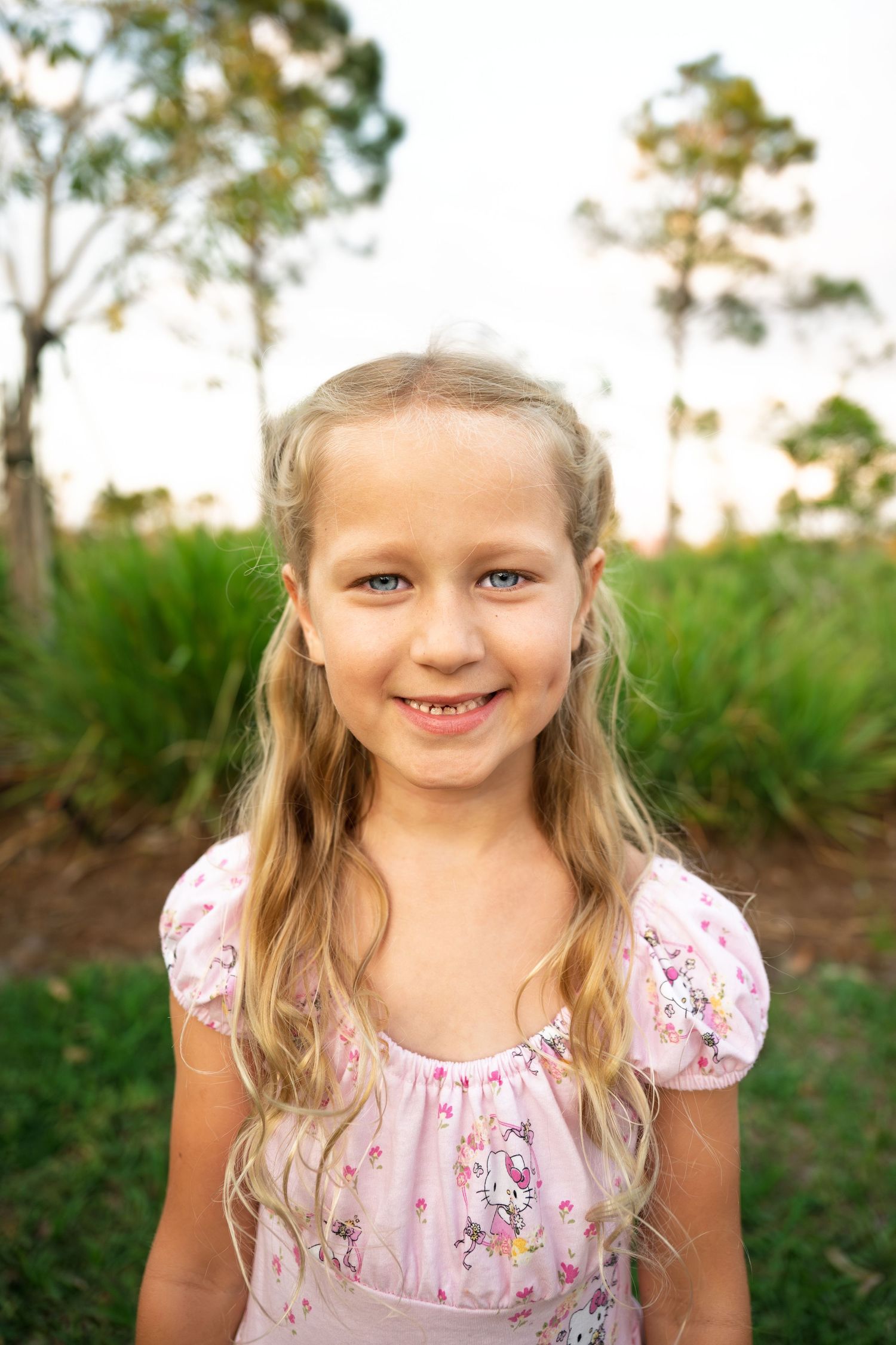 Smiling young blonde girl in a pink floral dress posing outdoors in a garden setting.