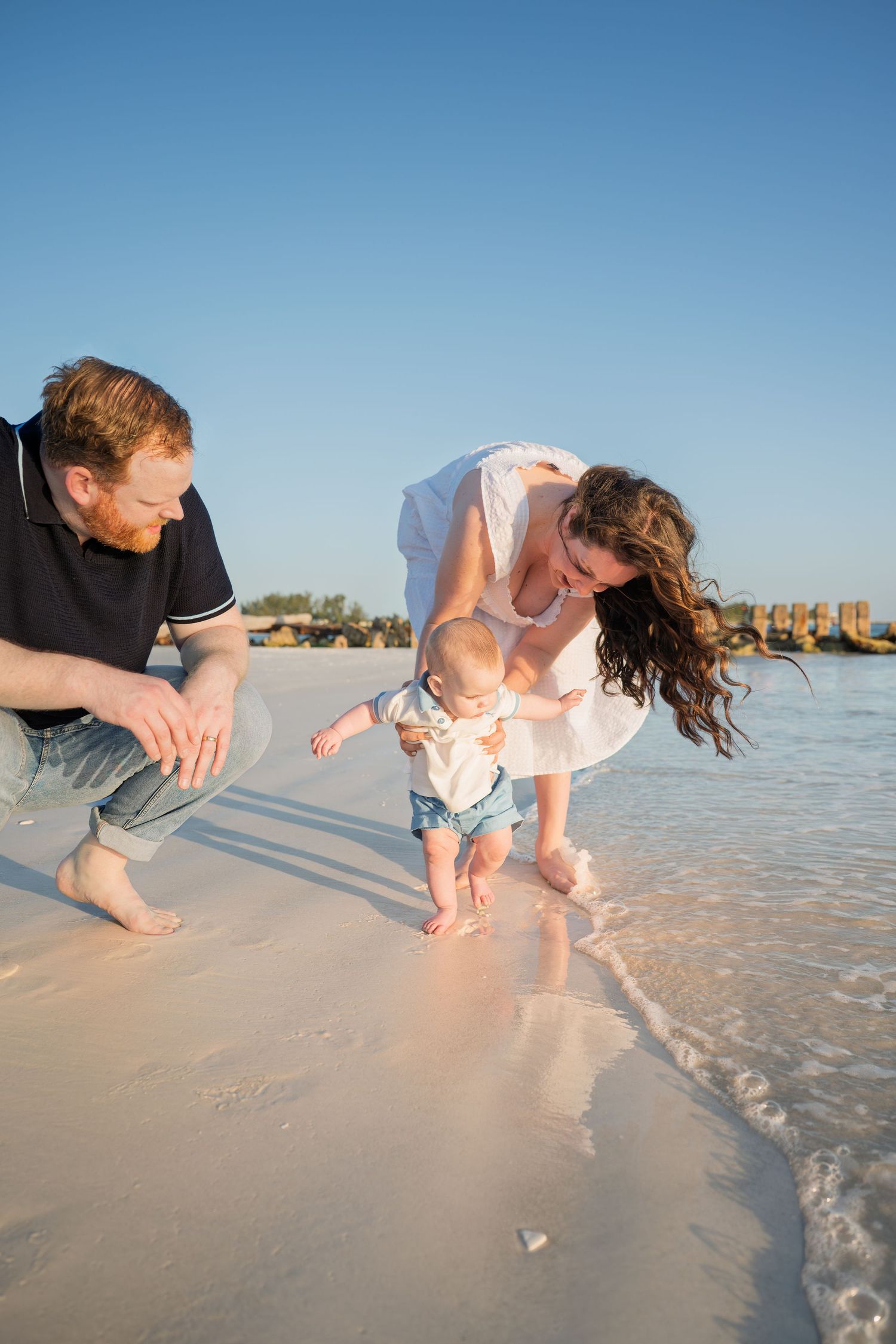 Young family helping baby take first steps on a sunny sandy beach with clear blue sky.