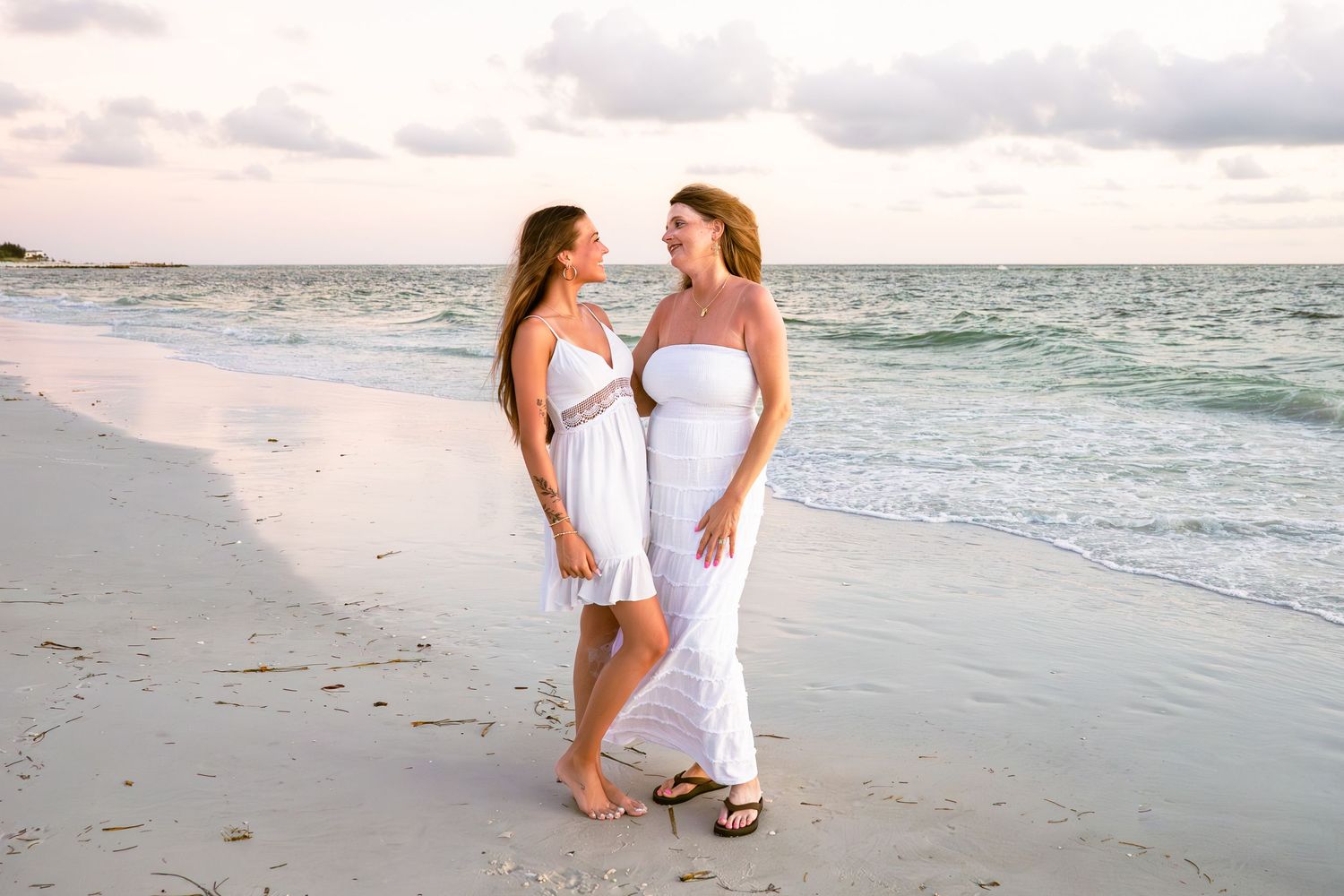 Two women in white dresses standing on a sandy beach at sunset, smiling at each other near the ocean waves.