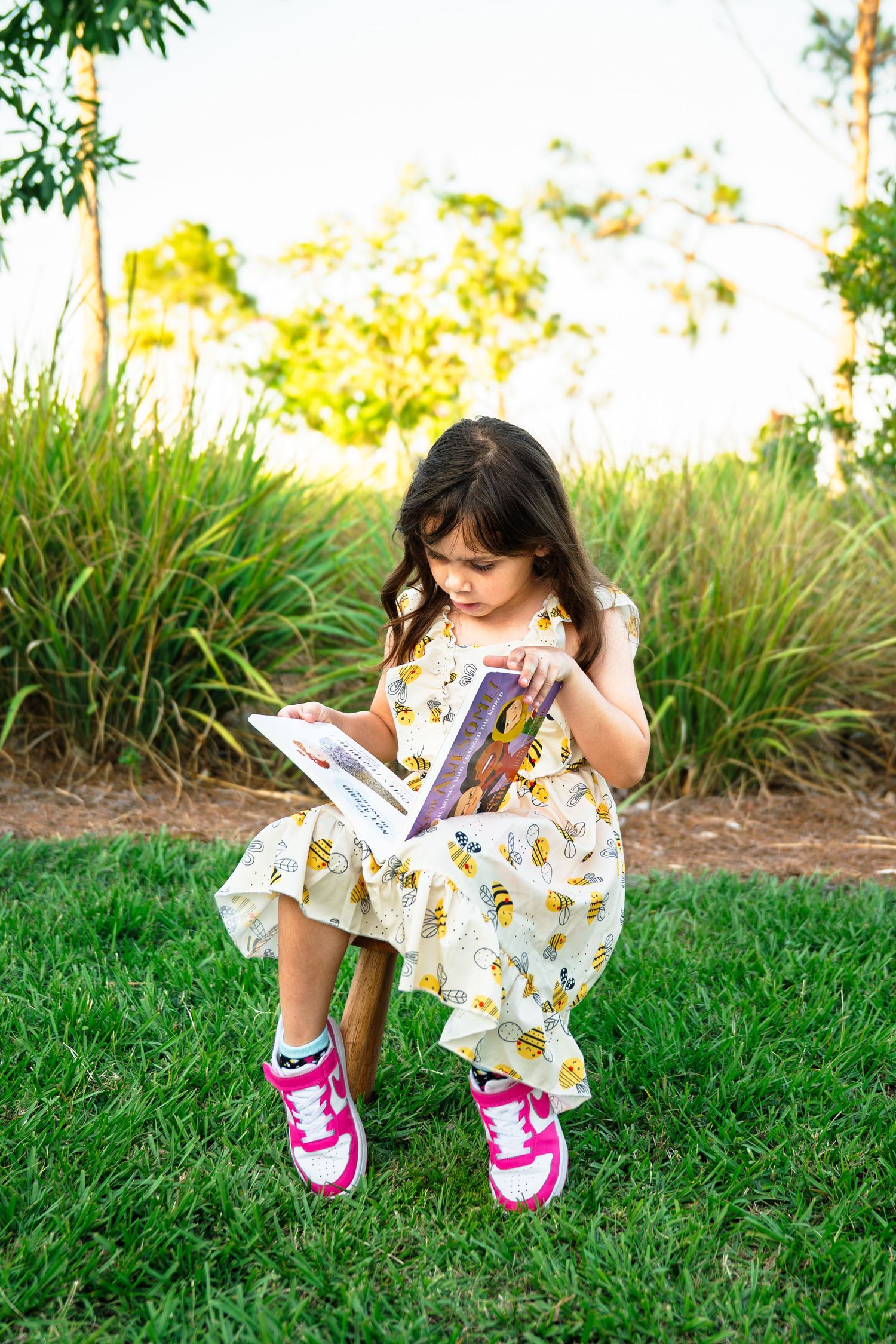 Young girl in bee-print dress and pink sneakers reading a book outdoors on green grass.