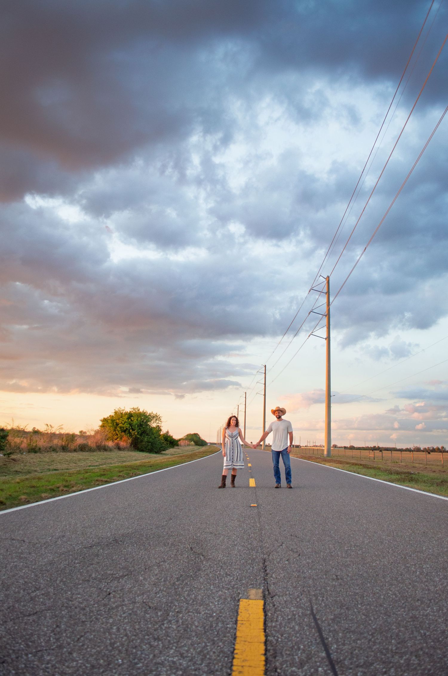 A couple and child hold hands while walking down an empty rural road under dramatic storm clouds at sunset.
