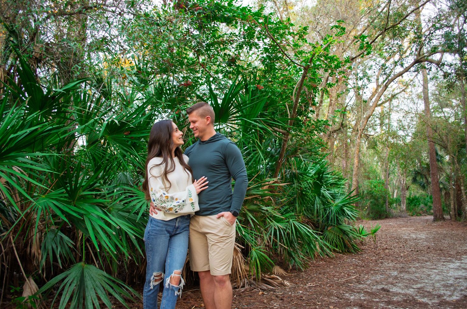Couple embraces on a romantic nature trail surrounded by lush tropical plants and palm trees.
