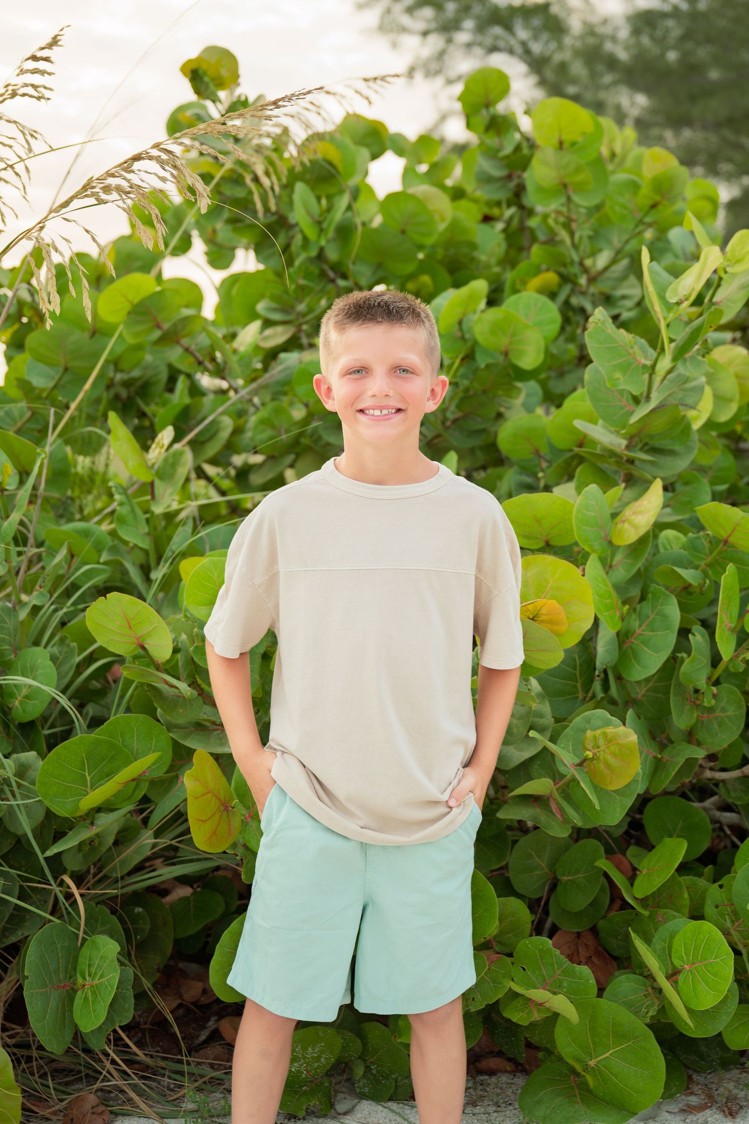 Smiling boy in beige shirt and mint shorts standing in front of lush green tropical foliage outdoors.