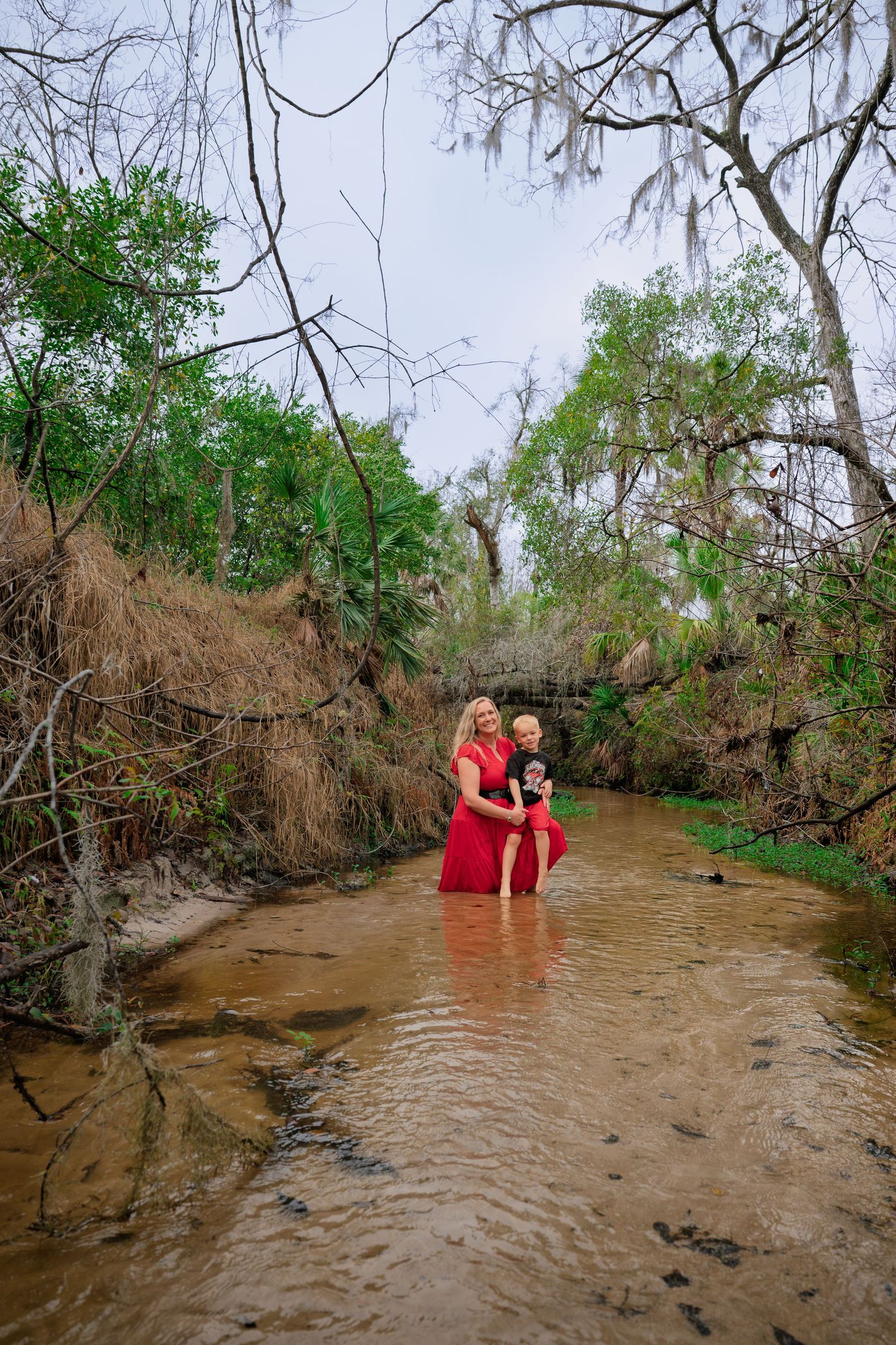 Woman in red dress holding child wading in a shallow creek surrounded by lush green trees.