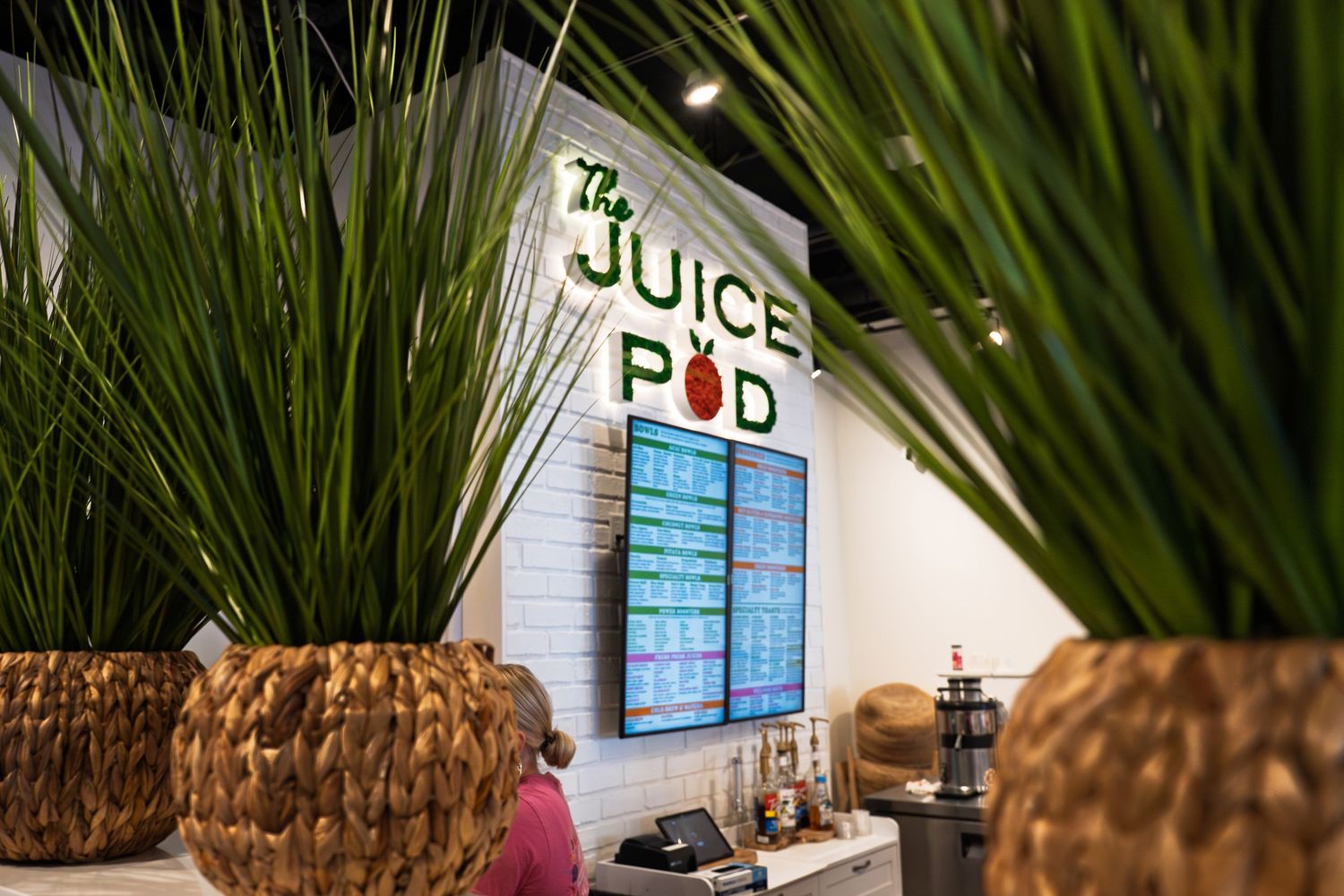 The Juice Pod restaurant interior with green plants in wicker baskets and a digital menu board on the wall.