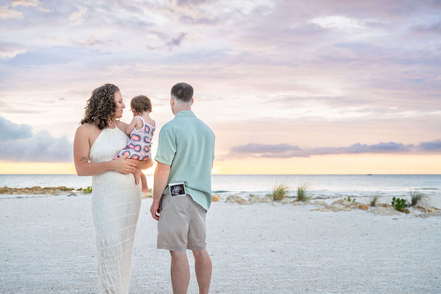 Family of three enjoying a beautiful pastel sunset on a white sand beach, parents holding toddler while gazing at the ocean.