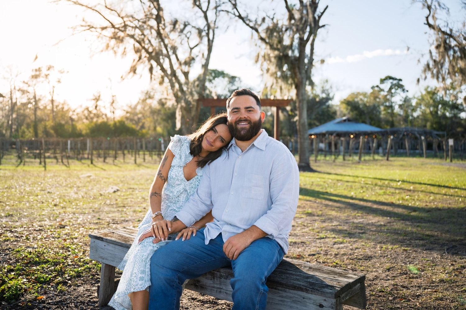 A couple shares a tender moment while sitting on a wooden bench in a sunlit park setting.