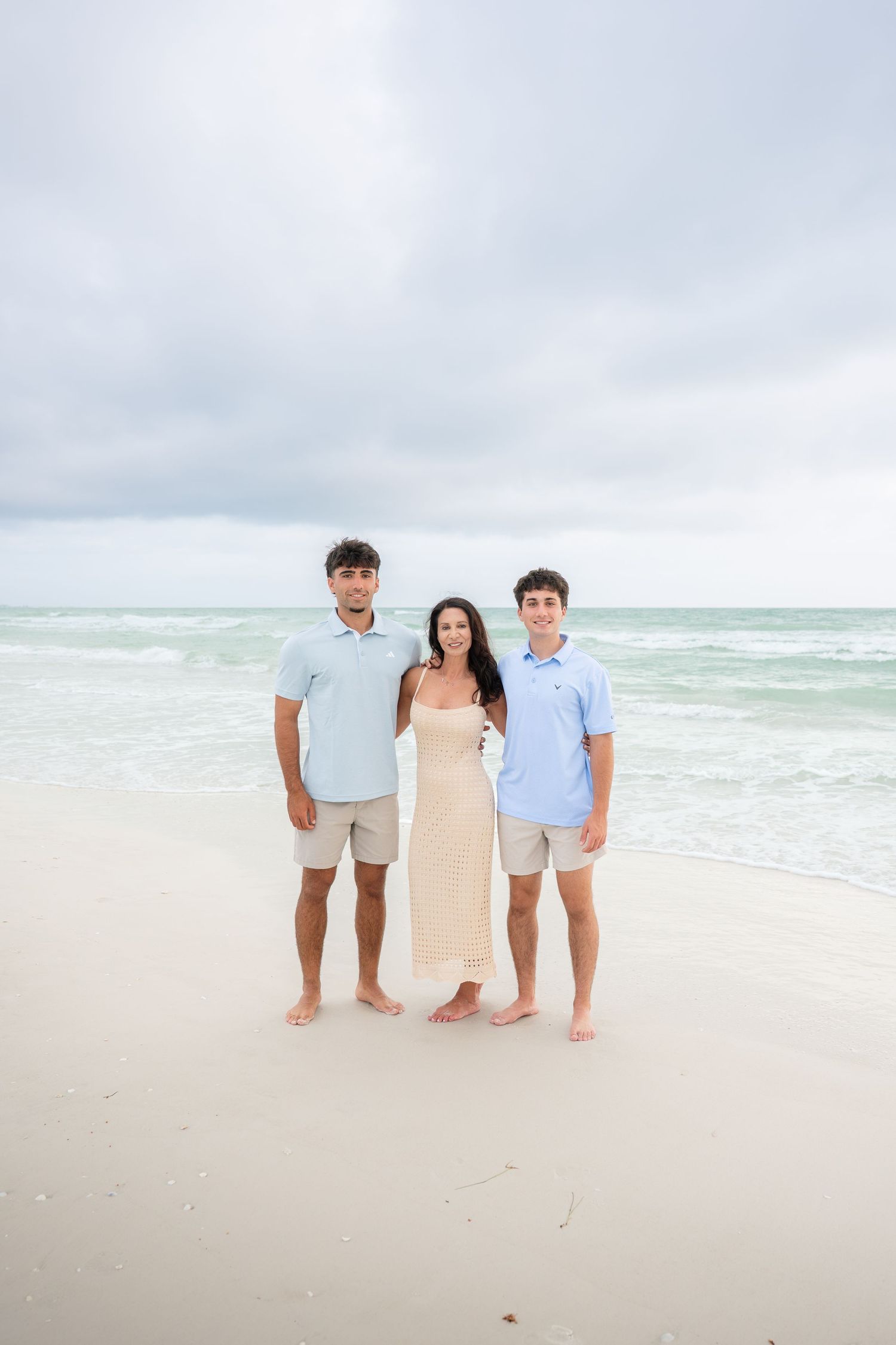 A woman and two teenage boys pose together on a white sandy beach with turquoise ocean waves behind them.
