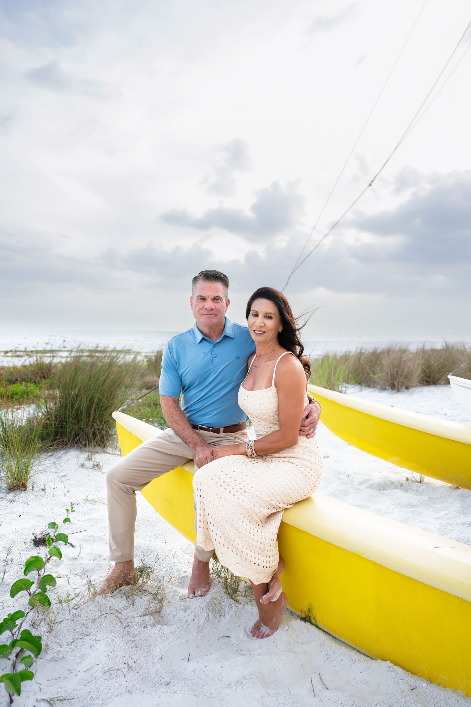 Couple sitting on a yellow boat on white sand beach, man in blue polo and woman in cream dress, cloudy sky backdrop.