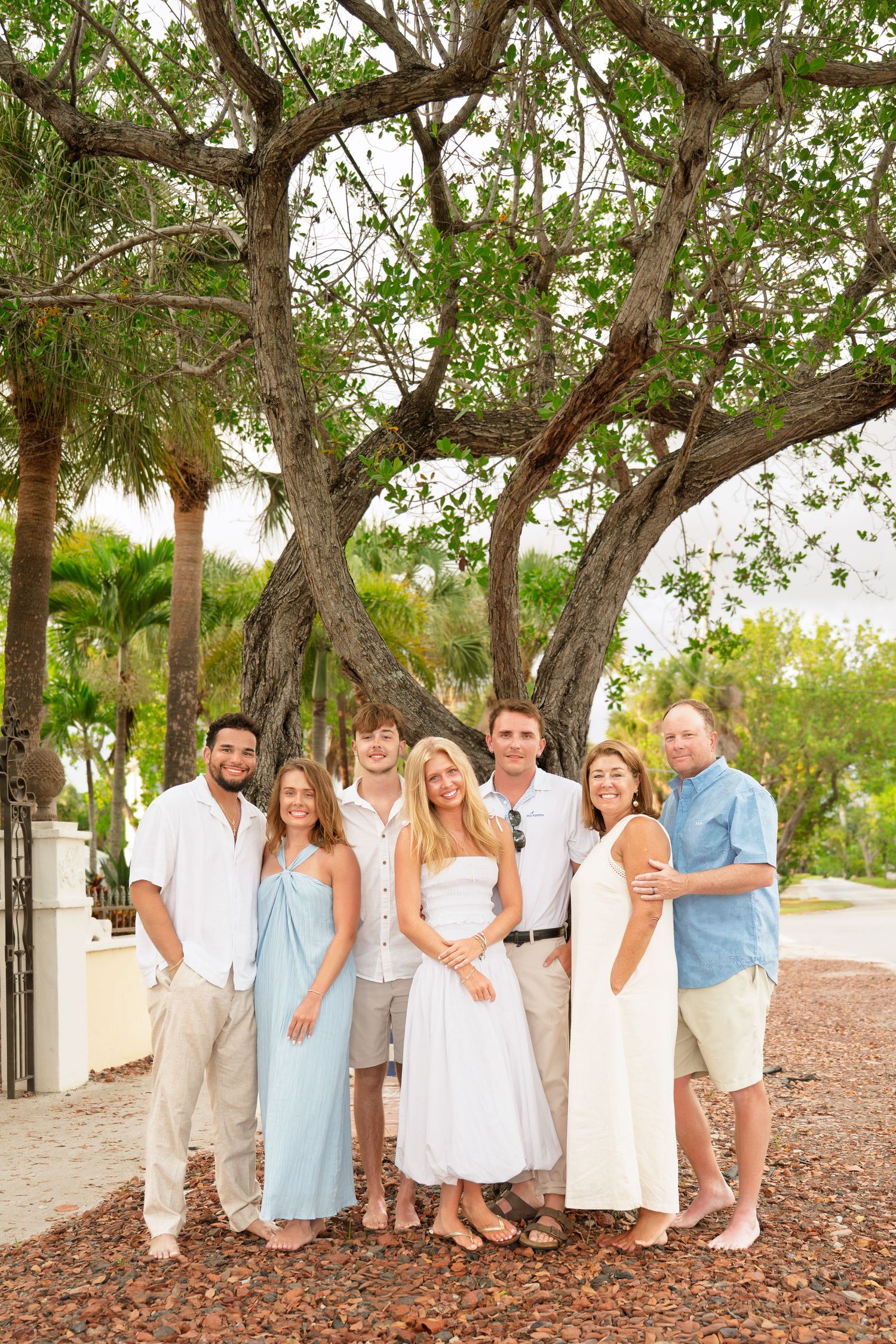 A happy family of eight poses together outdoors under a large tree, dressed in white and light blue casual attire.