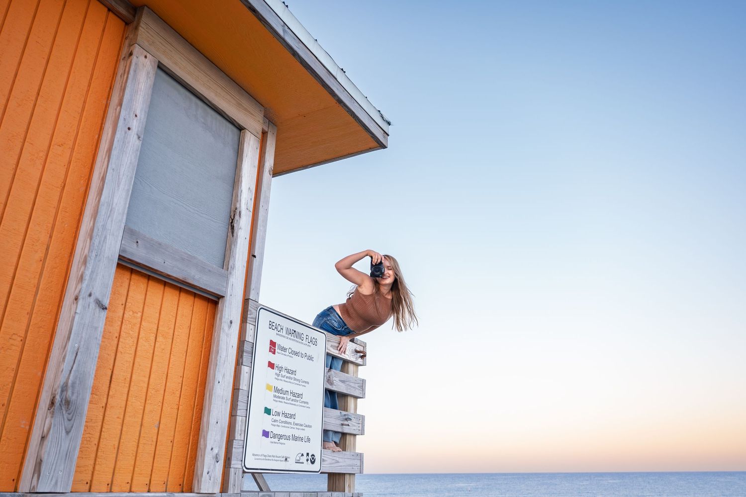 Woman sitting atop an orange lifeguard station sign at a beach during golden hour sunset.