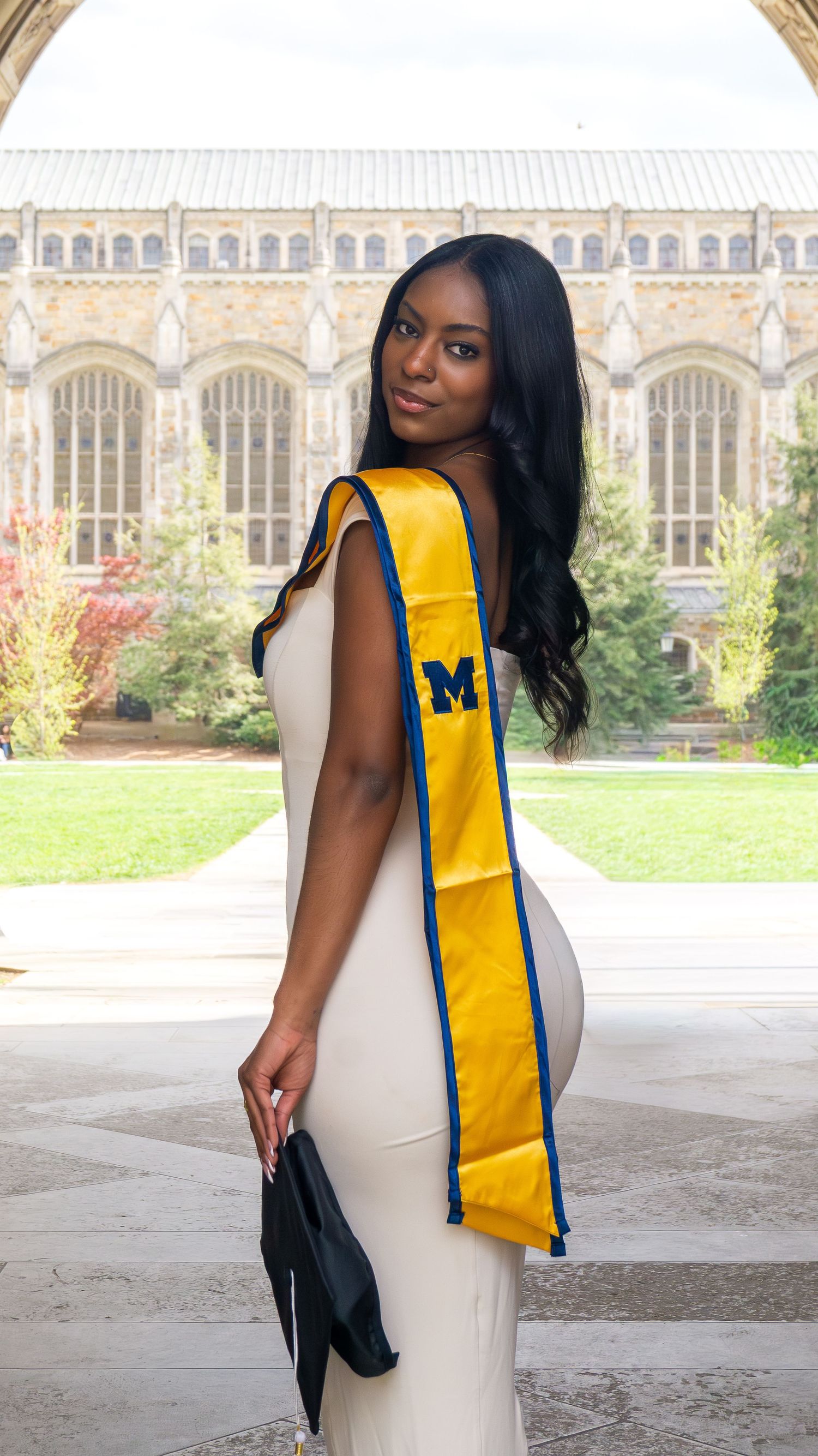 Young woman wearing University of Michigan gold graduation sash poses in a courtyard.