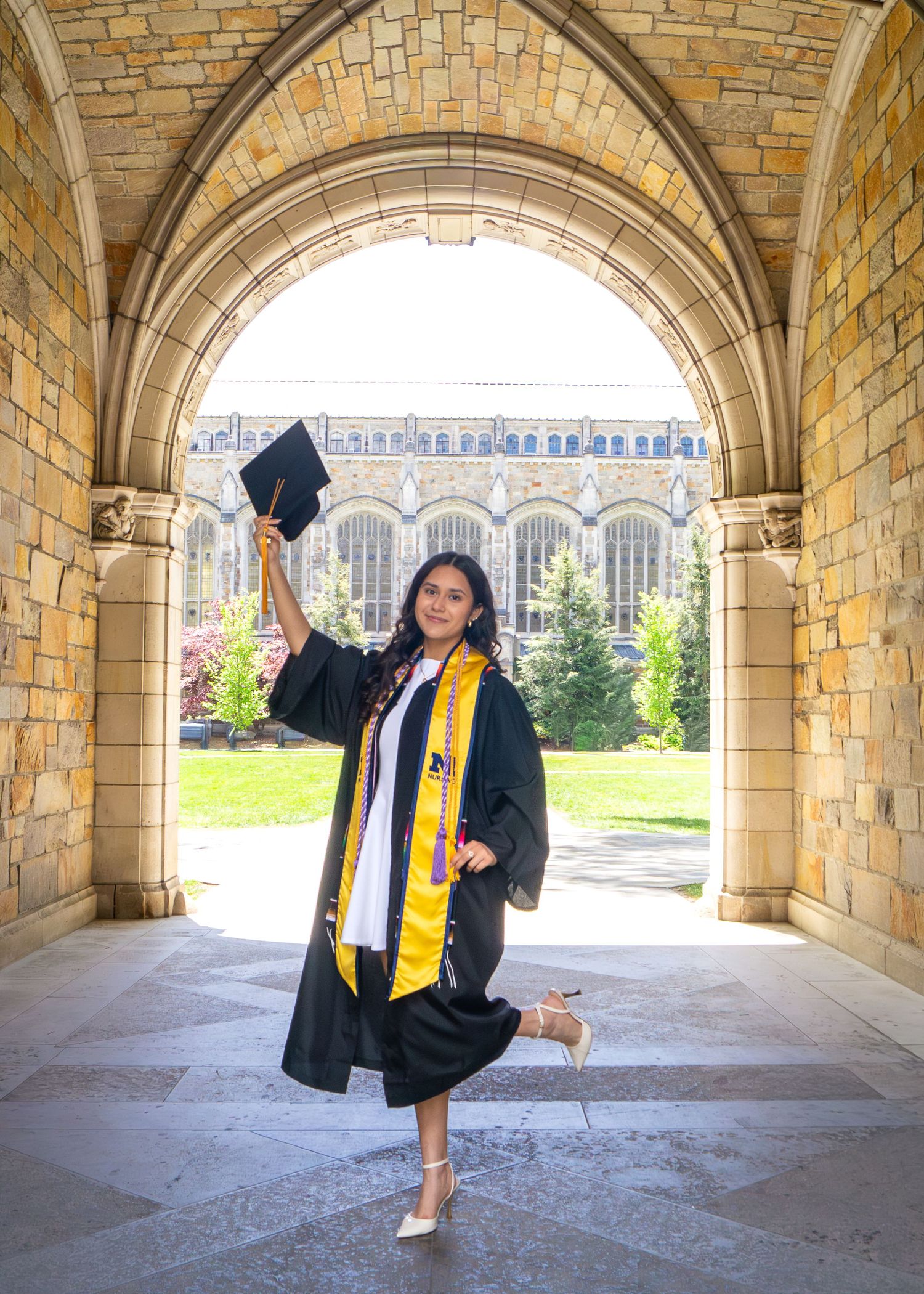 Graduate in black cap and gown with gold stole celebrates under stone archway on university campus.