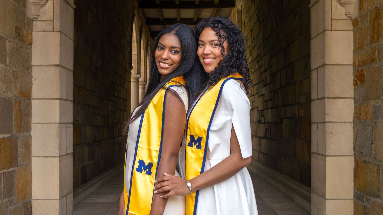 Two women in white outfits wearing University of Michigan graduation stoles pose back-to-back under a stone archway.