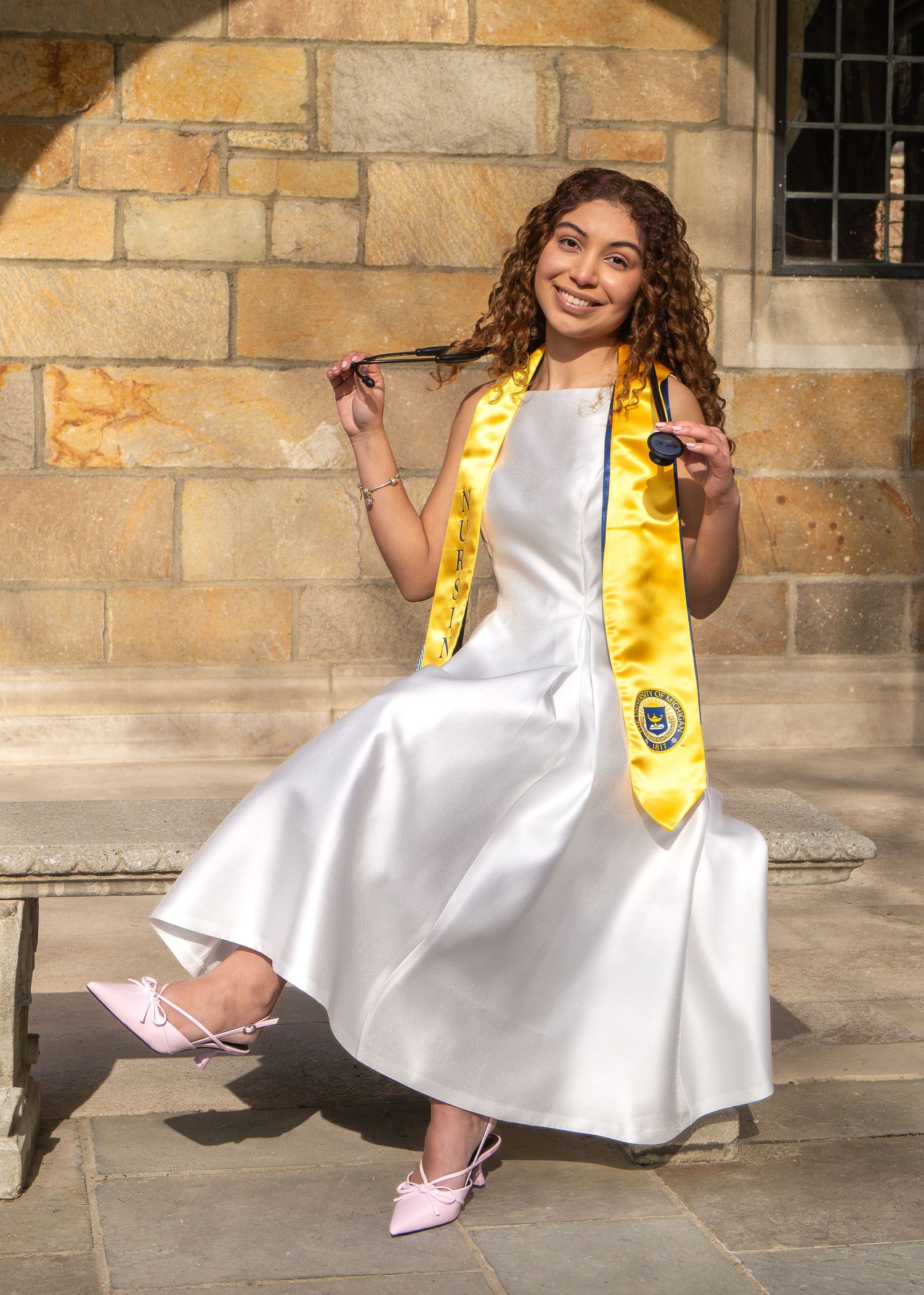 Young woman in white graduation dress and gold honor stole poses proudly on stone steps holding her cap tassel.