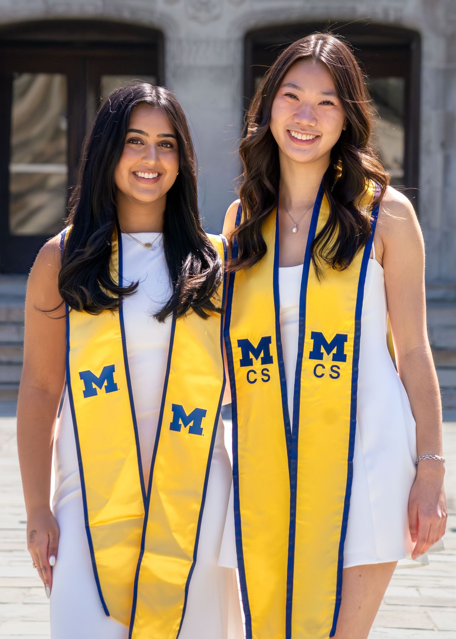 Two smiling University of Michigan graduates wearing gold M and CS stoles pose outdoors in white dresses on graduation day.