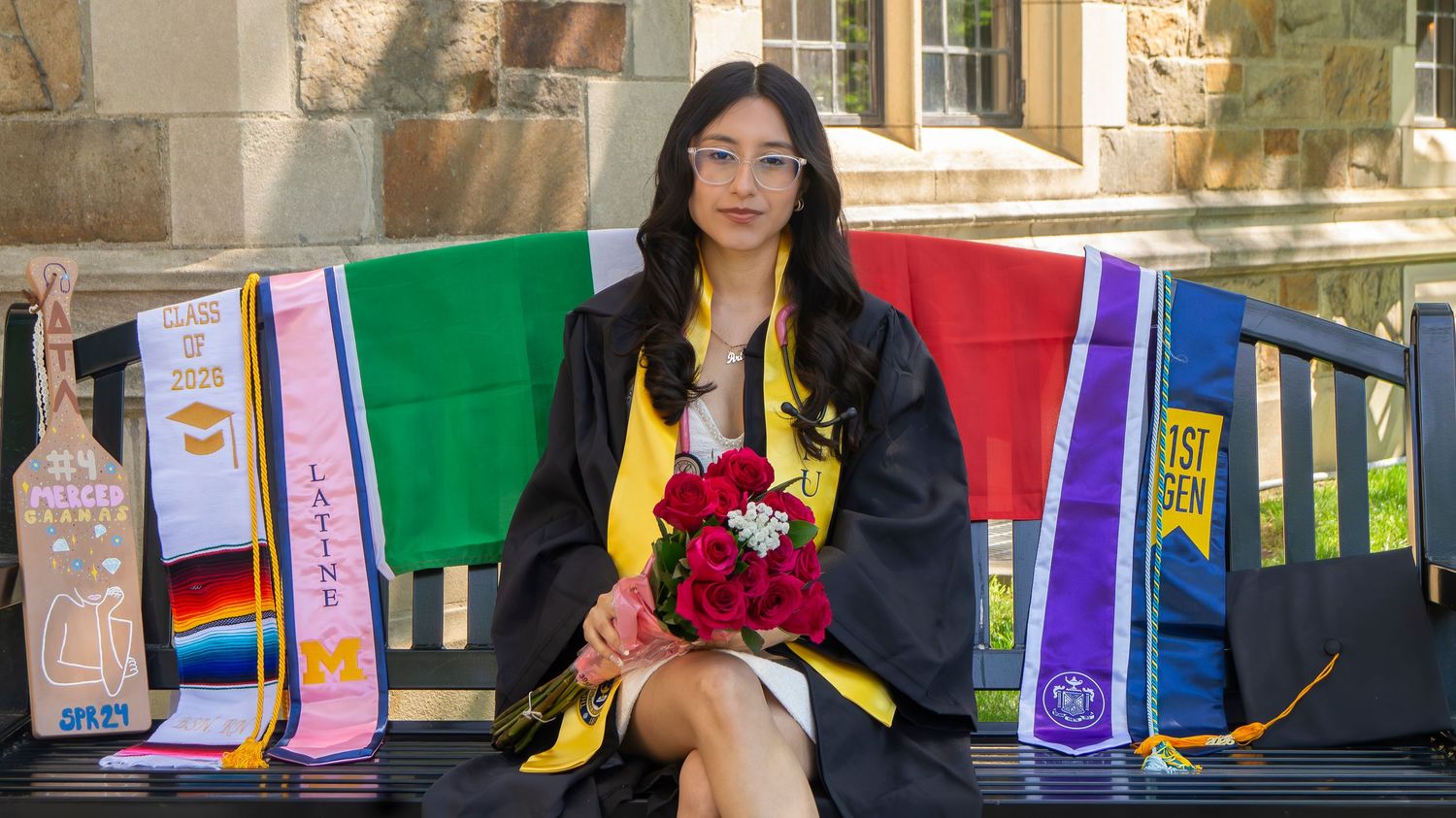 Graduate in cap and gown holds red roses, seated on a bench surrounded by colorful sashes at an outdoor ceremony.
