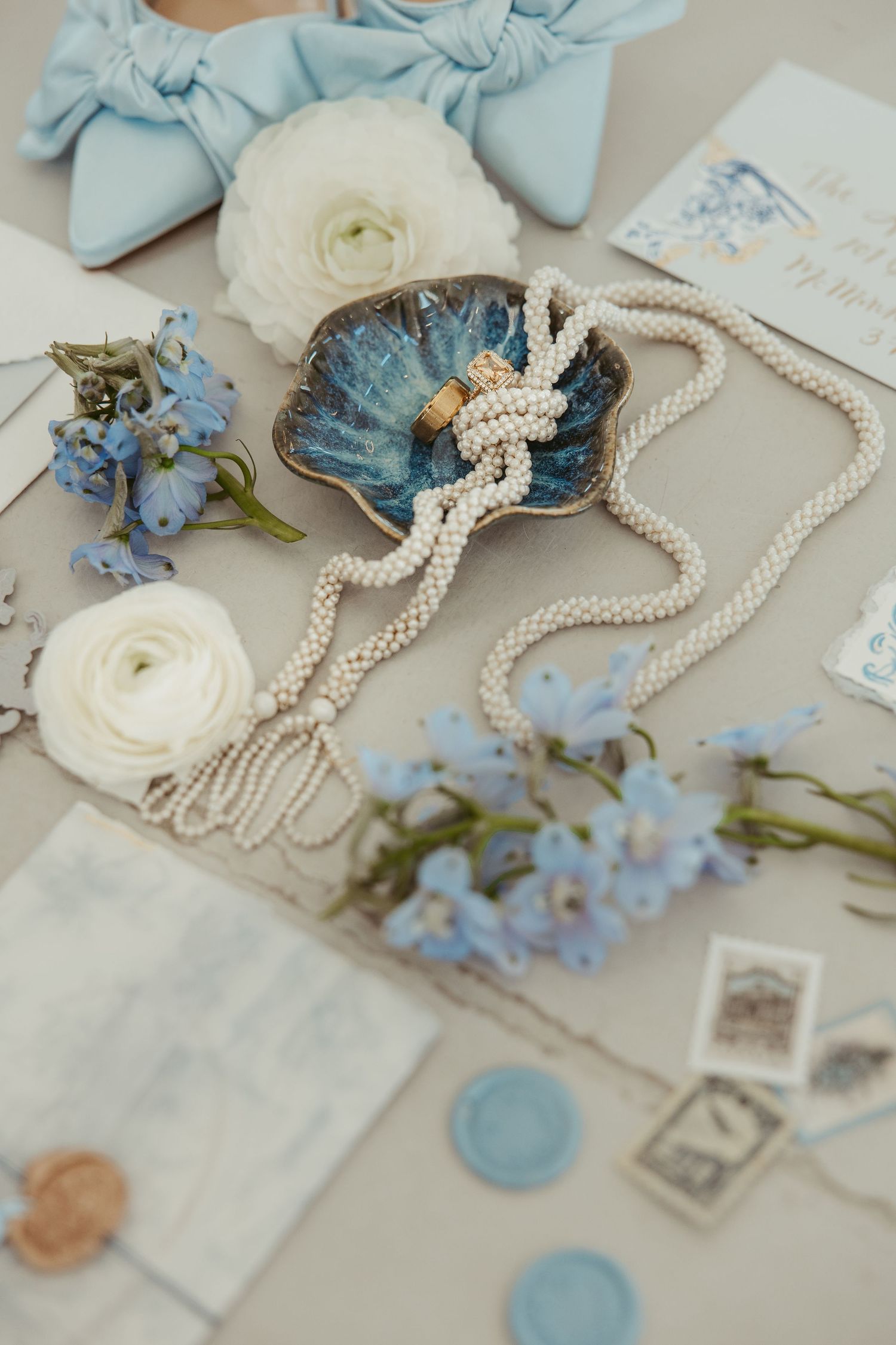 Wedding detail flat lay featuring heirloom rings, pearl jewelry, blue florals, bridal shoes, and romantic wedding stationery.