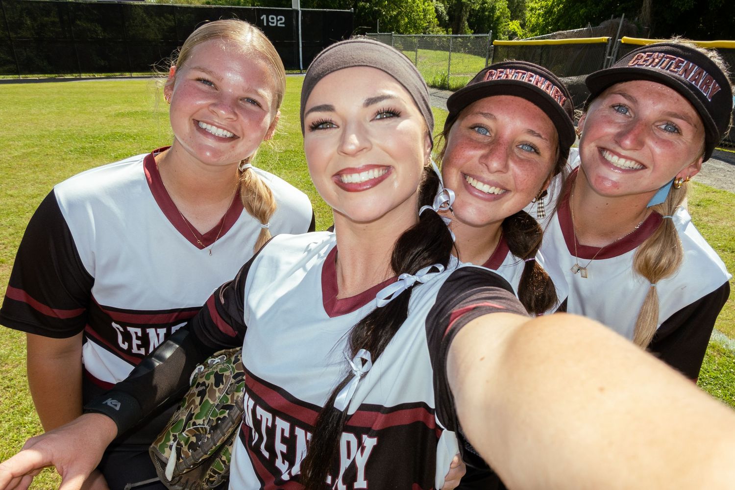 Four smiling female softball players in uniforms taking a selfie together outdoors.
