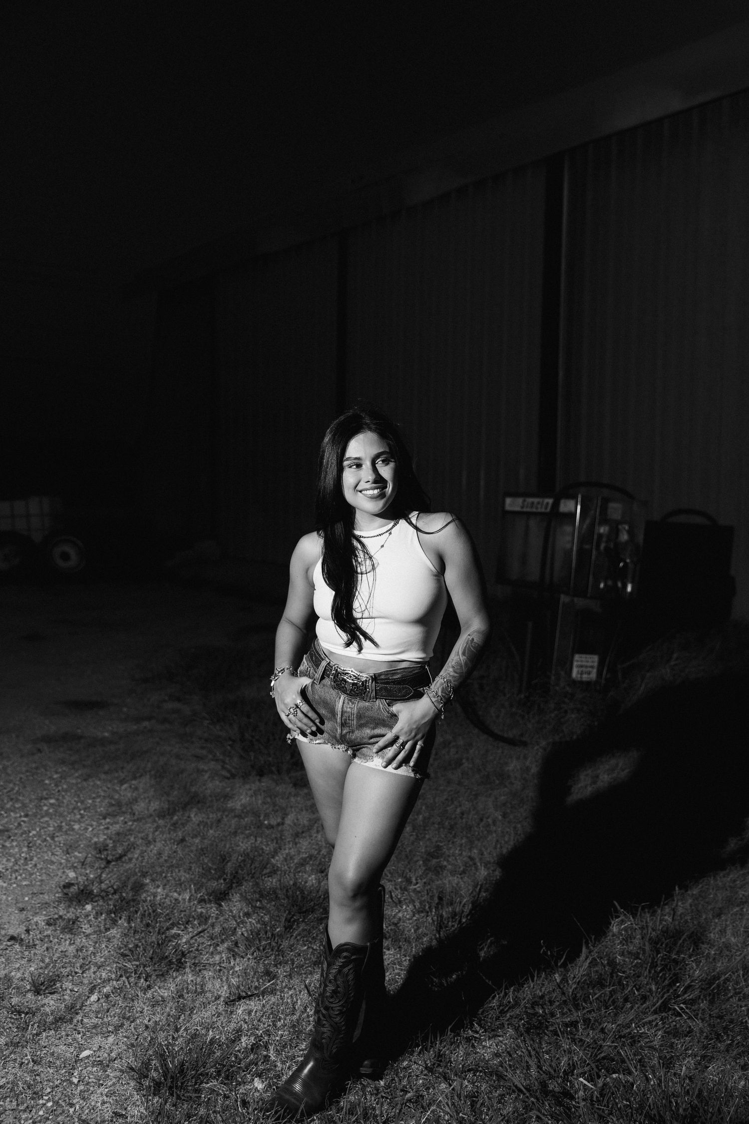 Black and white photo of a smiling young woman in shorts and boots posing outdoors at night near a barn.