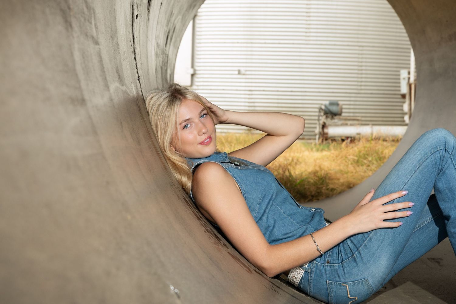 Blonde woman in denim jumpsuit posing inside a large concrete pipe outdoors.