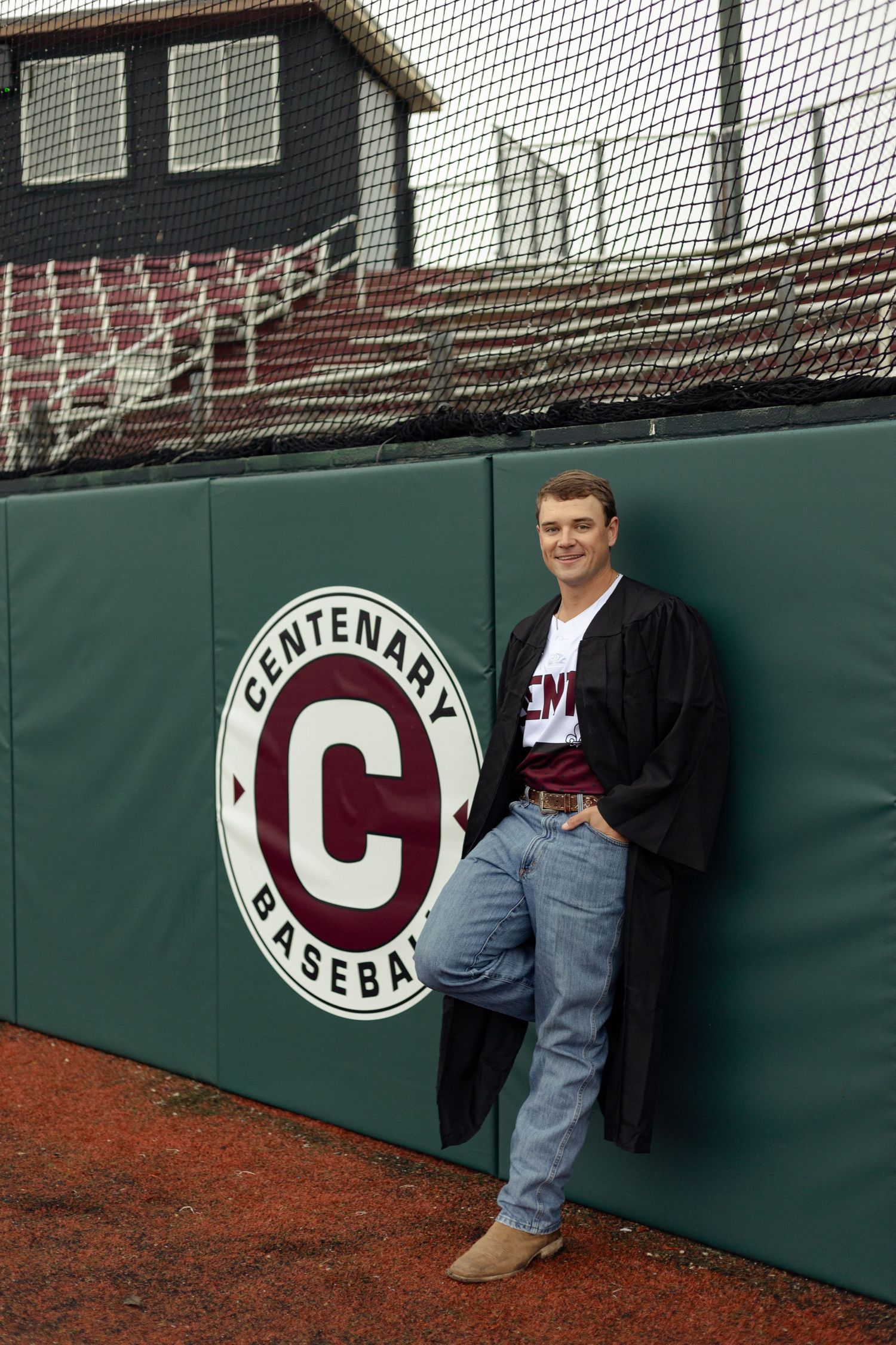 Young man in graduation gown leaning against Centenary Baseball outfield wall at a baseball field.
