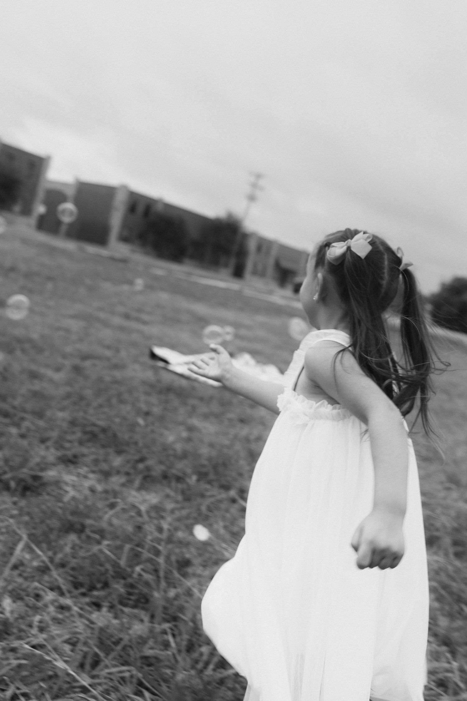 Black and white photo of a young girl in a white dress chasing bubbles in a grassy field.