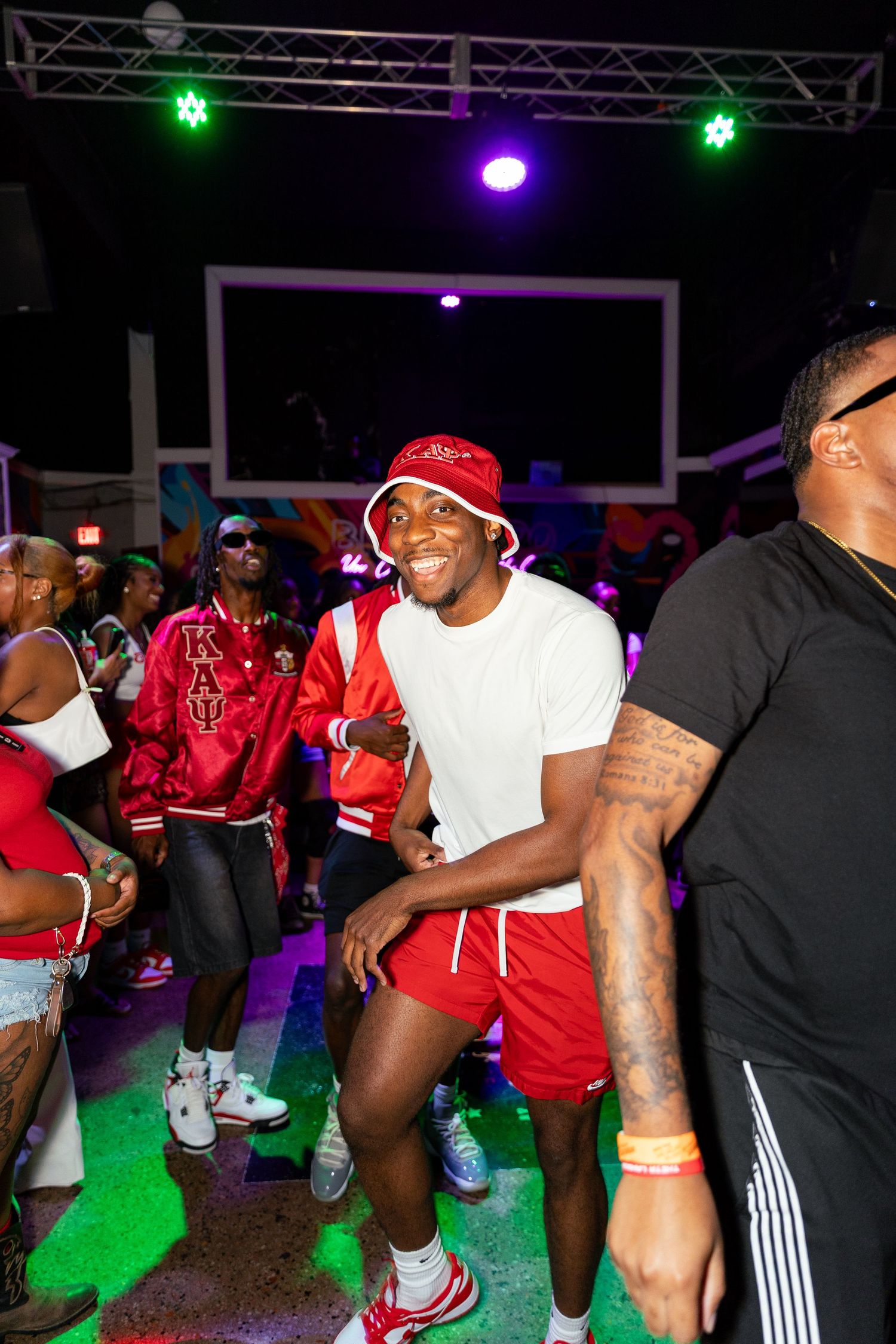 Young man in white outfit and red bucket hat dancing at a vibrant nightclub party with colorful lights.