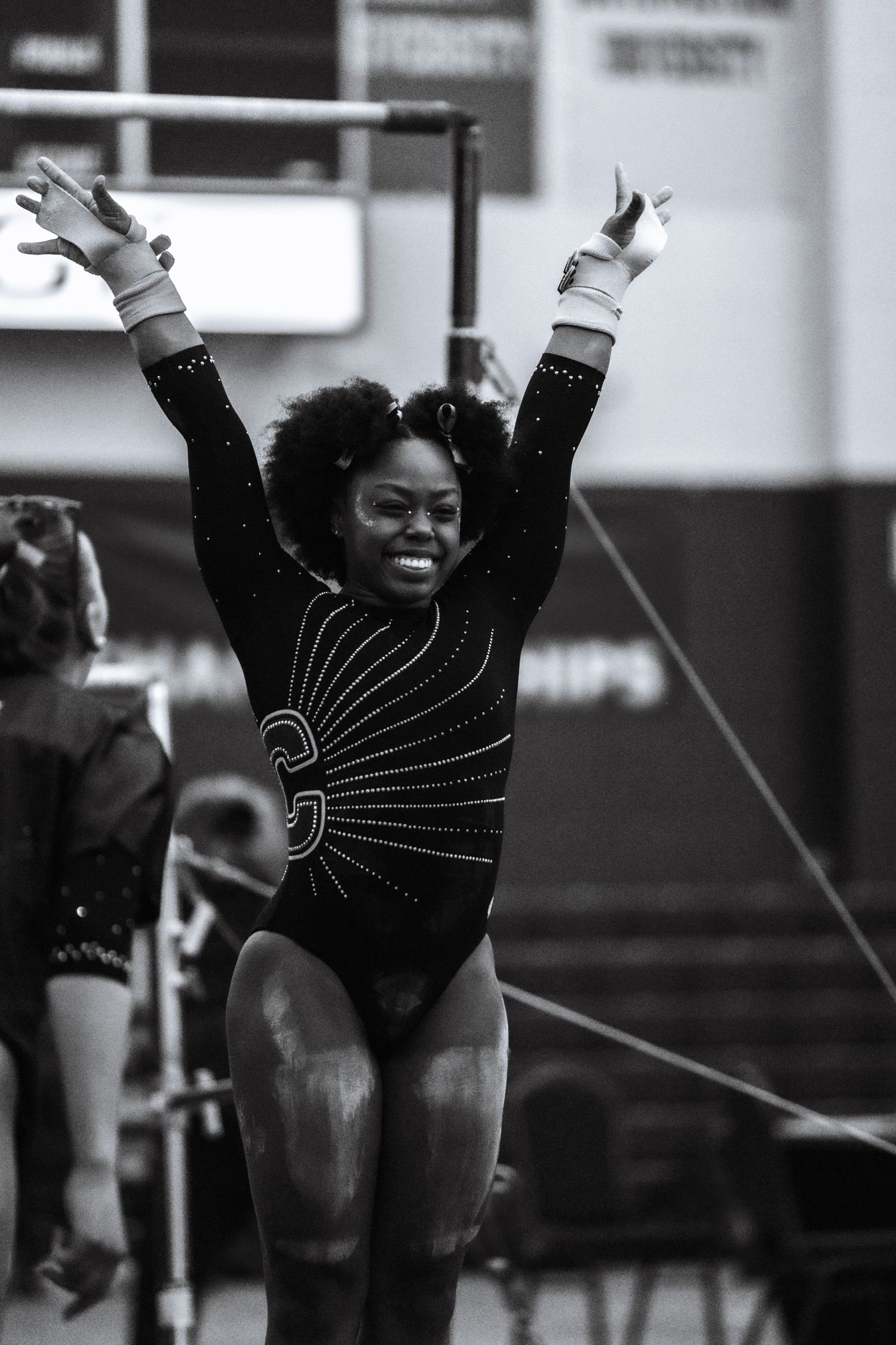 Female gymnast celebrating with arms raised in black sparkle leotard after routine, black and white action photo.
