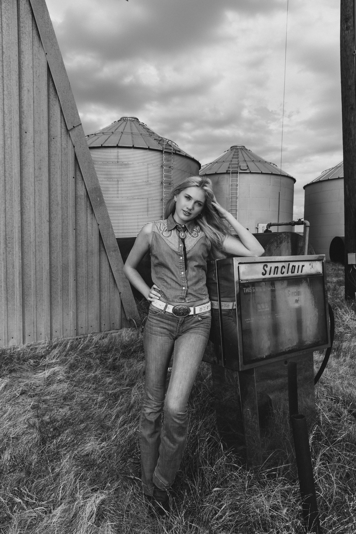 Black and white photo of a young woman in cowboy boots and jeans posing by a Sinclair gas pump with grain silos behind her.