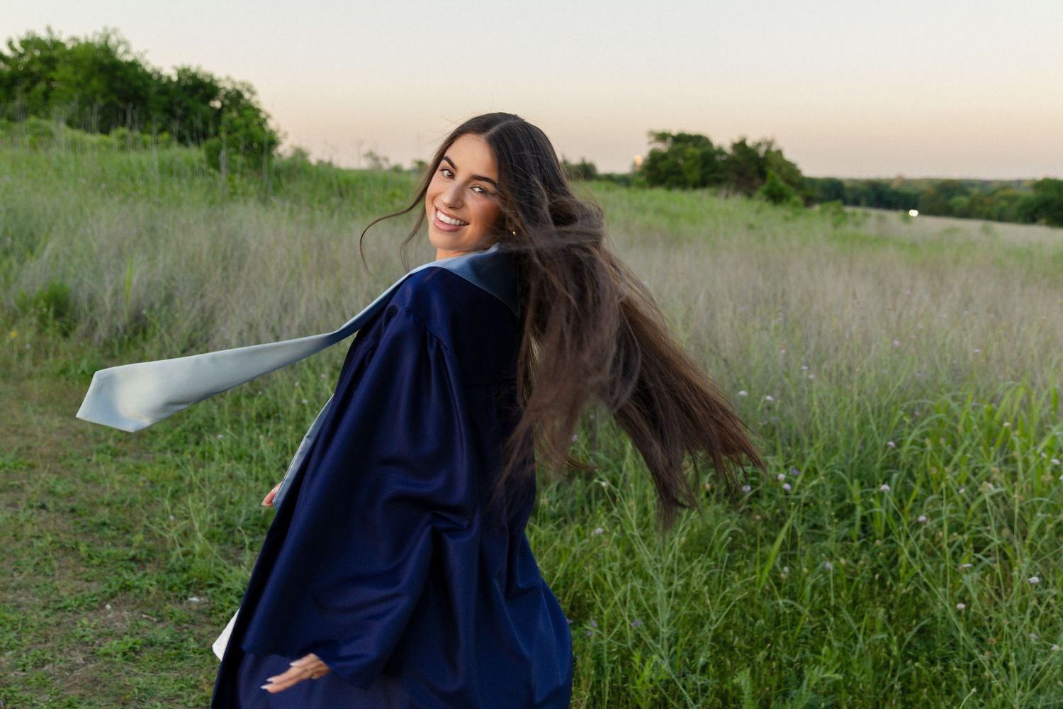 Smiling young woman in navy blue graduation gown twirling in a lush green field at sunset.