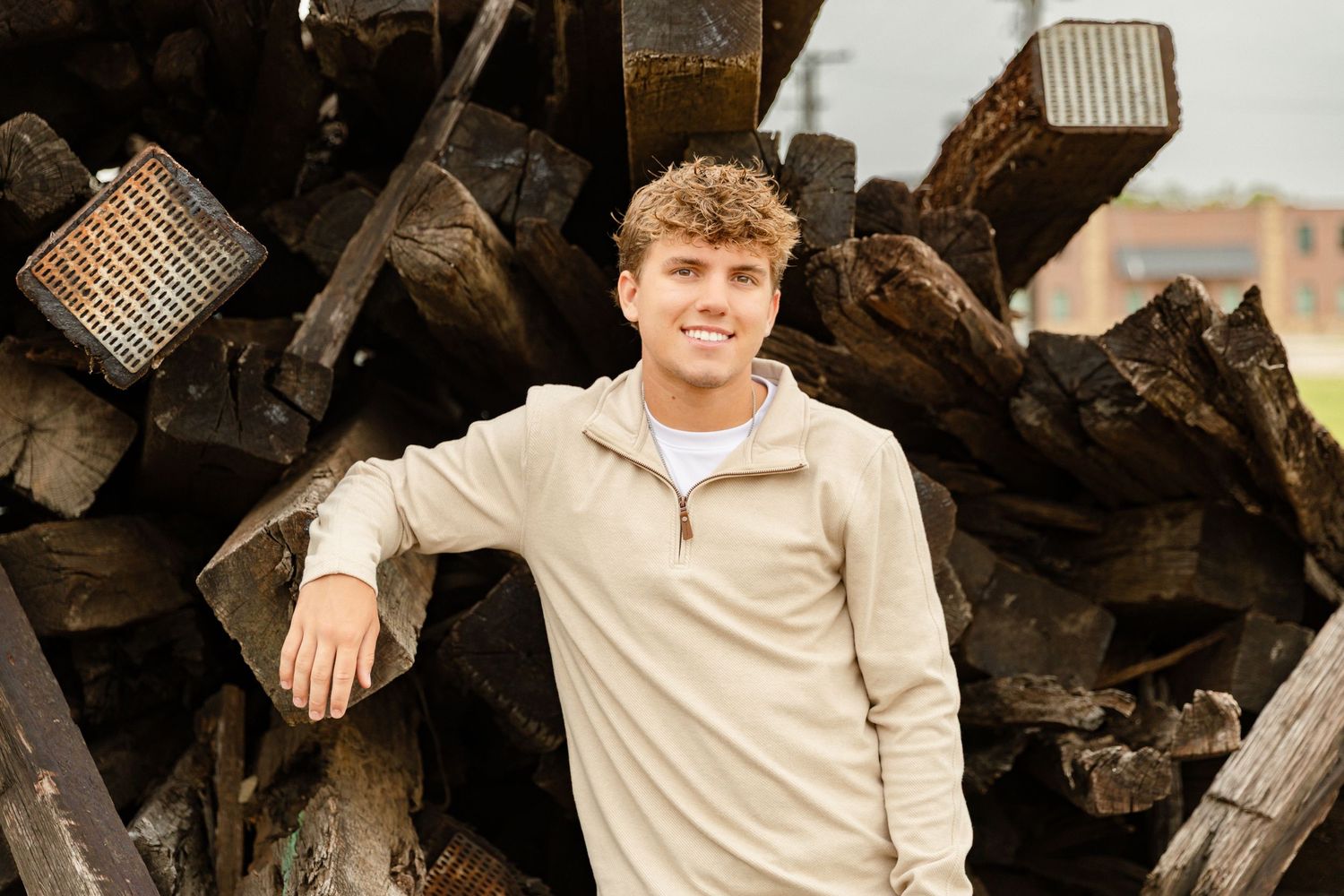 Young man in beige pullover smiling outdoors, leaning against a stack of rustic dark wooden logs.