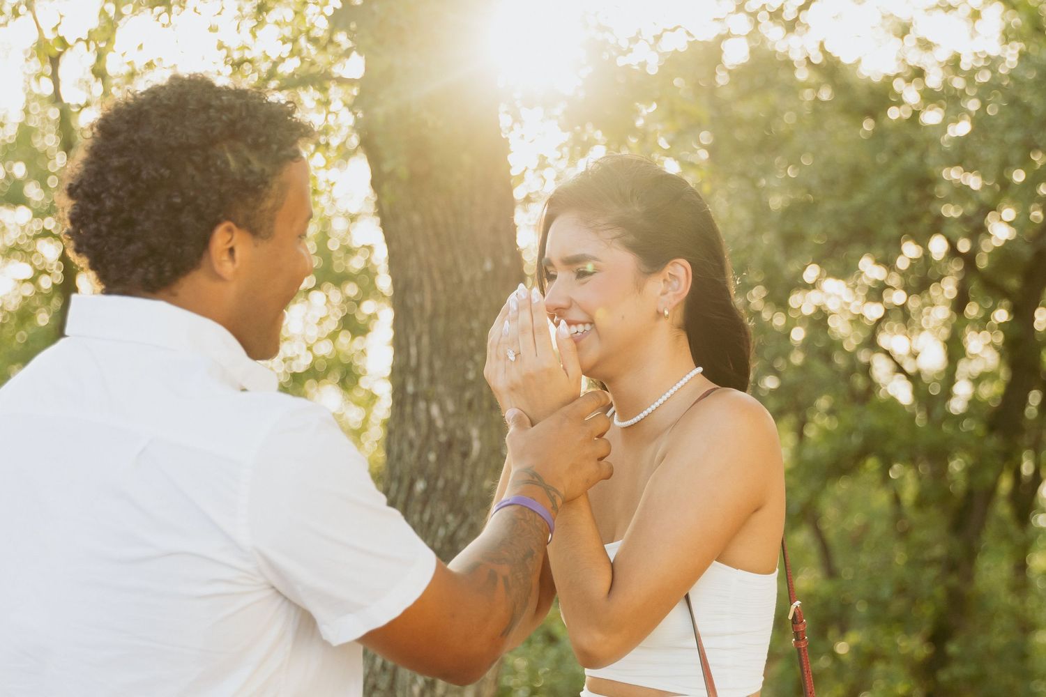 Couple sharing a romantic proposal moment outdoors in golden sunlight, woman covering mouth in surprise.