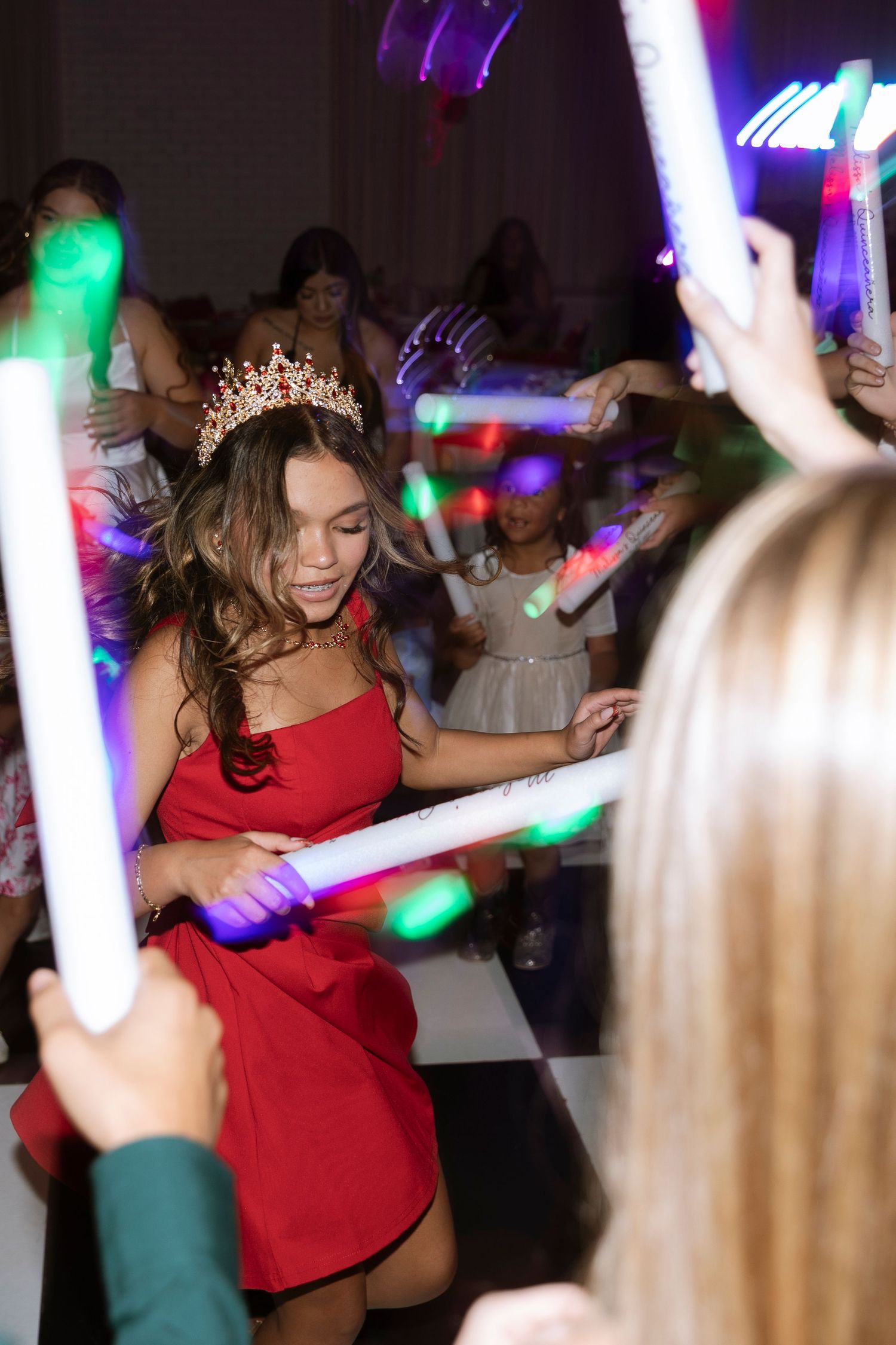 Teen girl in red dress and tiara dances with glowing foam sticks at a quinceañera party celebration.