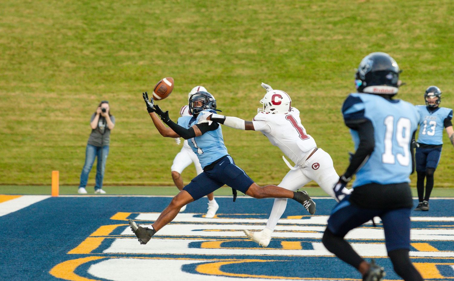 Wide receiver in blue jersey attempts catch in end zone during competitive football game.